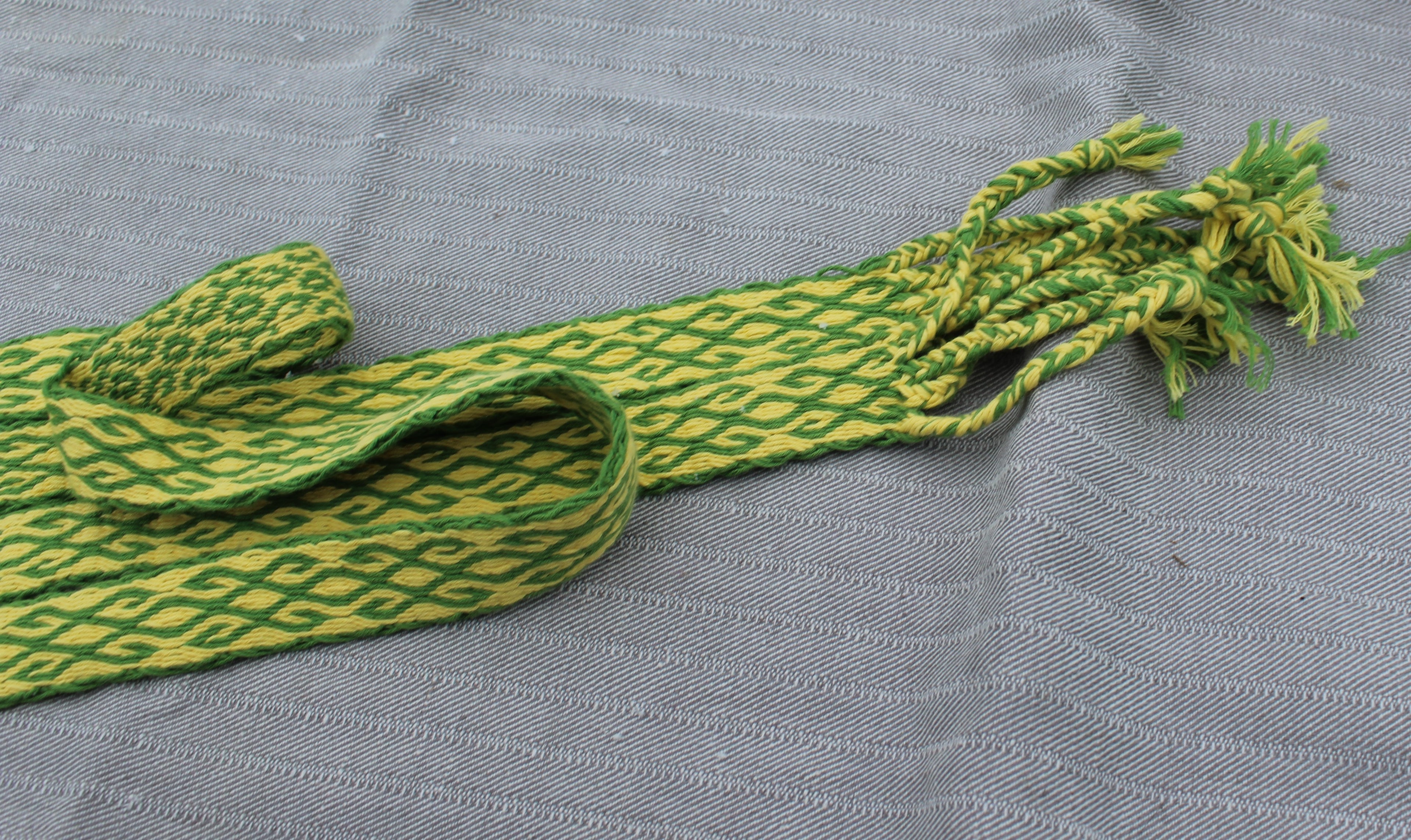 A green and yellow patterned woven strap bag with knotted tassels, laid flat on a textured gray fabric background.