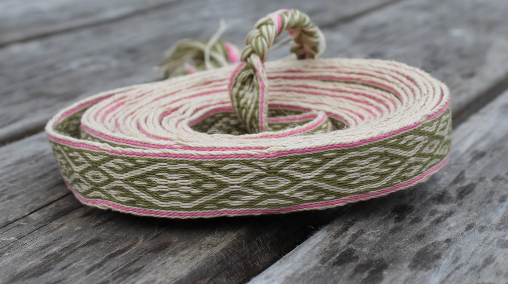 A coiled piece of woven cotton ribbon with a geometric pattern in olive green, cream, and pink, resting on a rustic gray wooden surface.