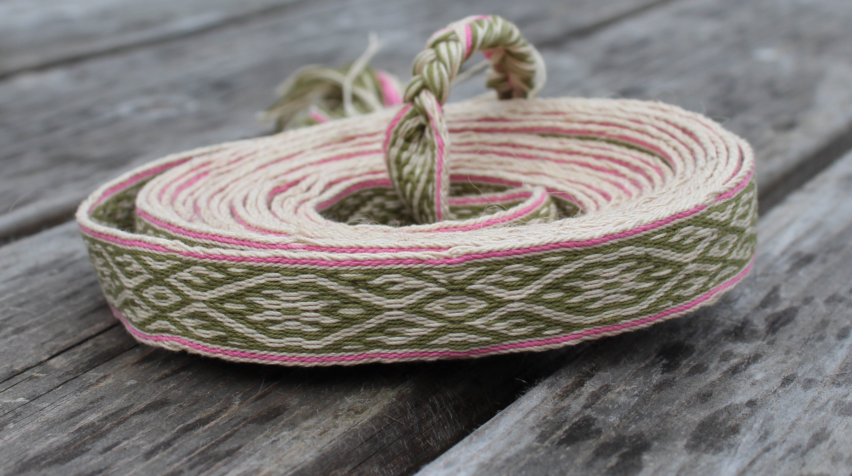 A coiled piece of woven cotton ribbon with a geometric pattern in olive green, cream, and pink, resting on a rustic gray wooden surface.