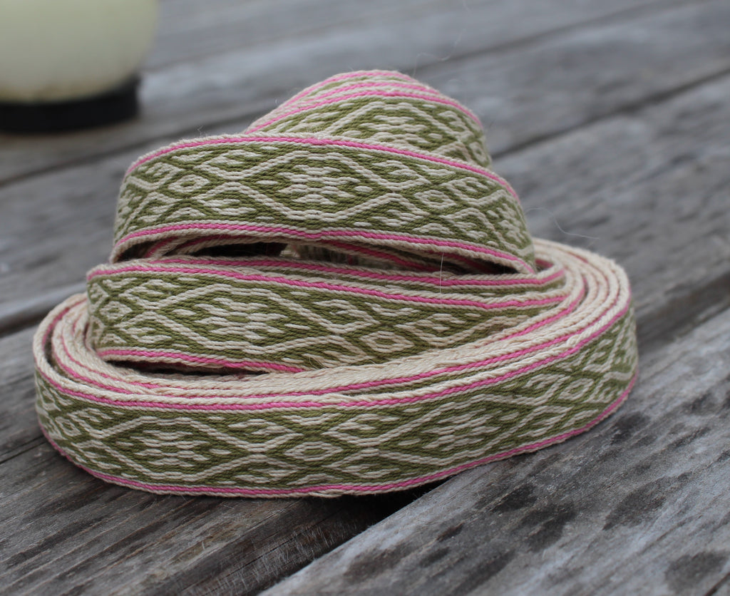 A coiled piece of woven ribbon trim with an olive green and cream geometric pattern and pink edging, displayed on a weathered gray wooden surface.