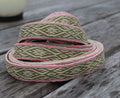 A coiled piece of woven ribbon trim with an olive green and cream geometric pattern and pink edging, displayed on a weathered gray wooden surface.