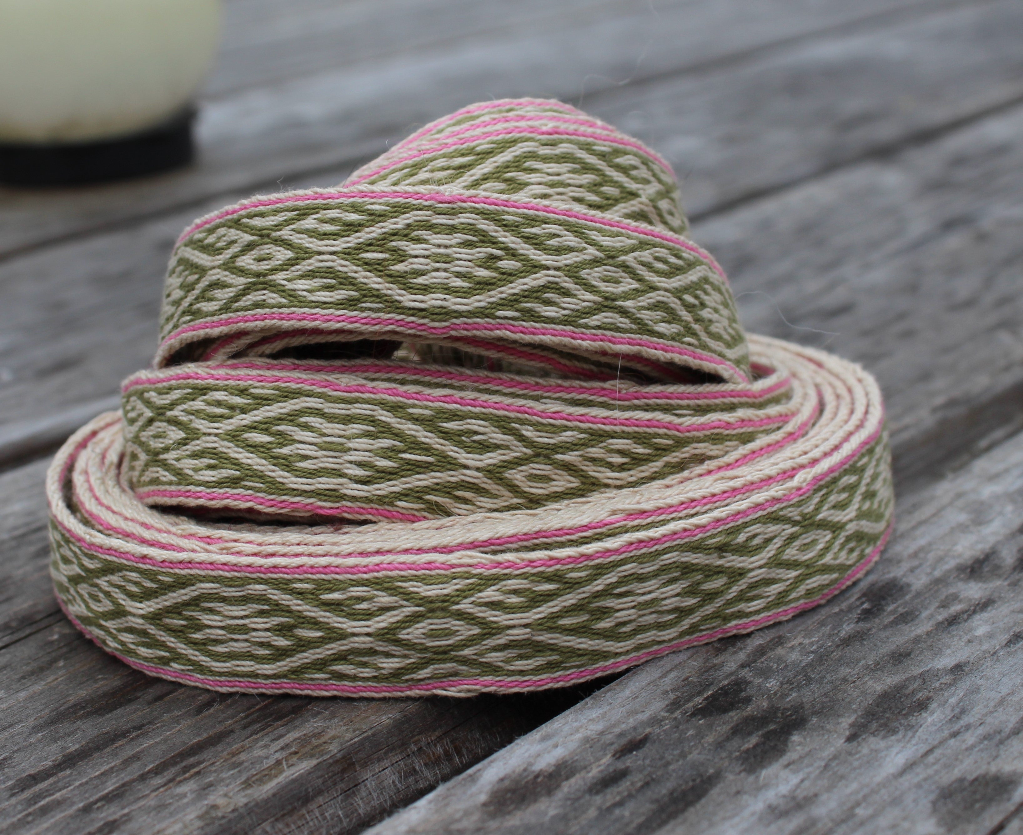 A coiled piece of woven ribbon trim with an olive green and cream geometric pattern and pink edging, displayed on a weathered gray wooden surface.
