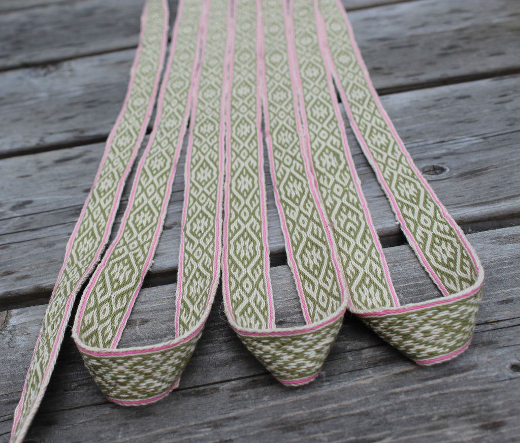 A close-up of a woven elastic strap with a green and white geometric diamond pattern and pink edging, laid out on a weathered gray wooden surface.