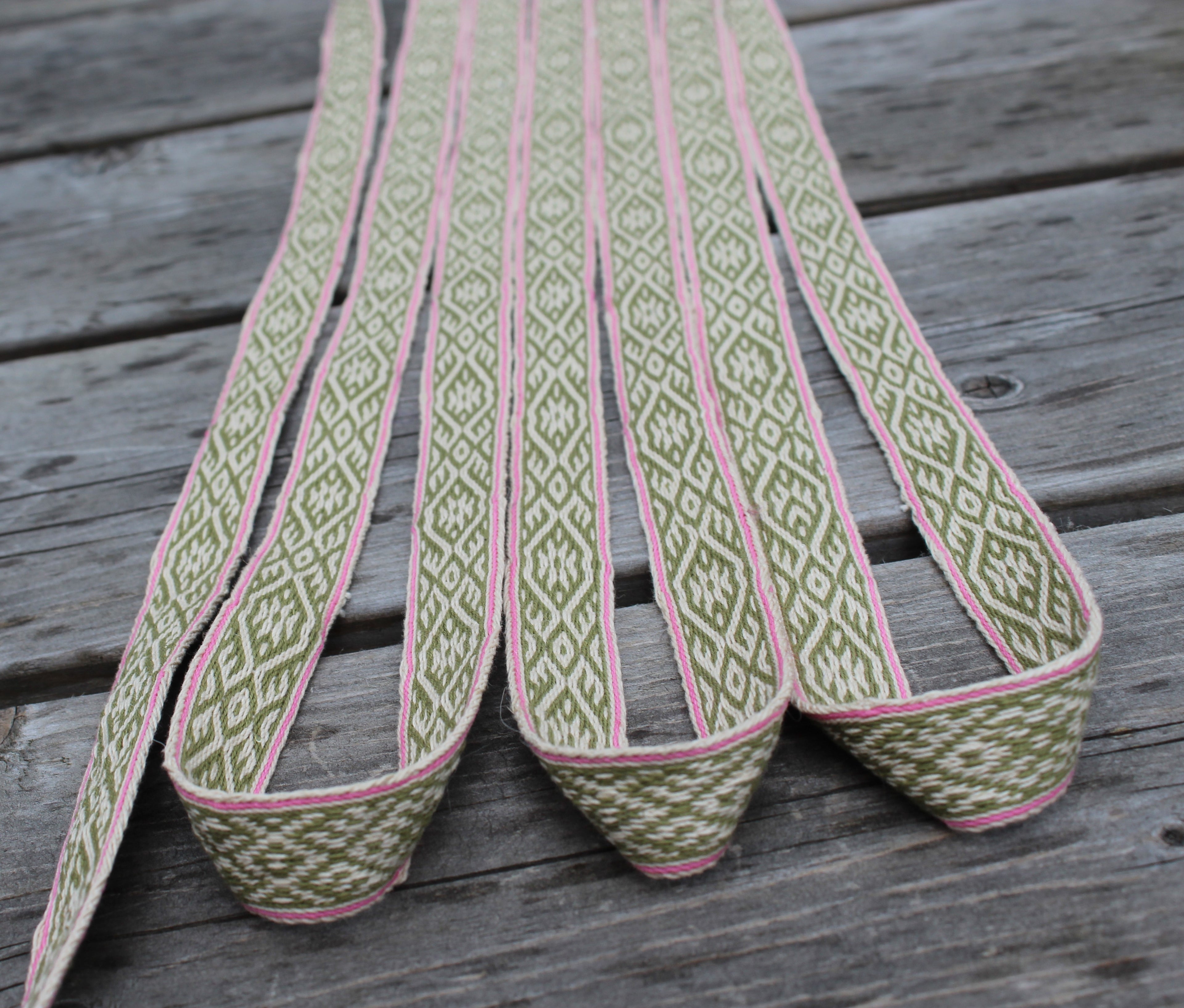 A close-up of a woven elastic strap with a green and white geometric diamond pattern and pink edging, laid out on a weathered gray wooden surface.
