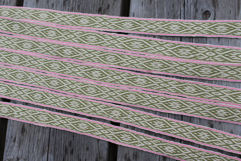 A close-up of multiple narrow woven elastic or ribbon strips laid diagonally on a weathered gray wooden surface. The fabric has an allover geometric diamond pattern in olive green and cream, bordered by thin pink stripes.