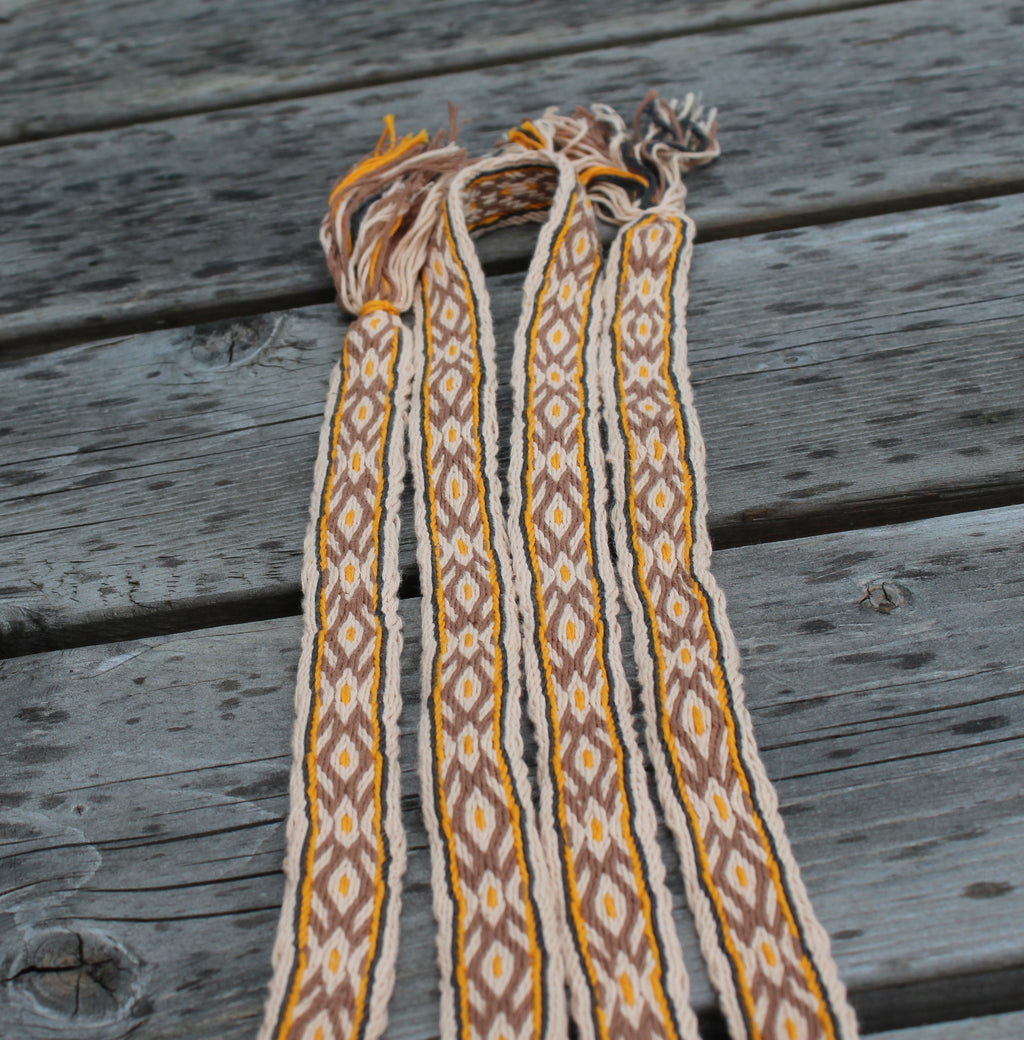 Three woven braided cords with fringe, featuring a geometric diamond pattern in brown, yellow, and cream, laid flat on weathered gray wooden planks.