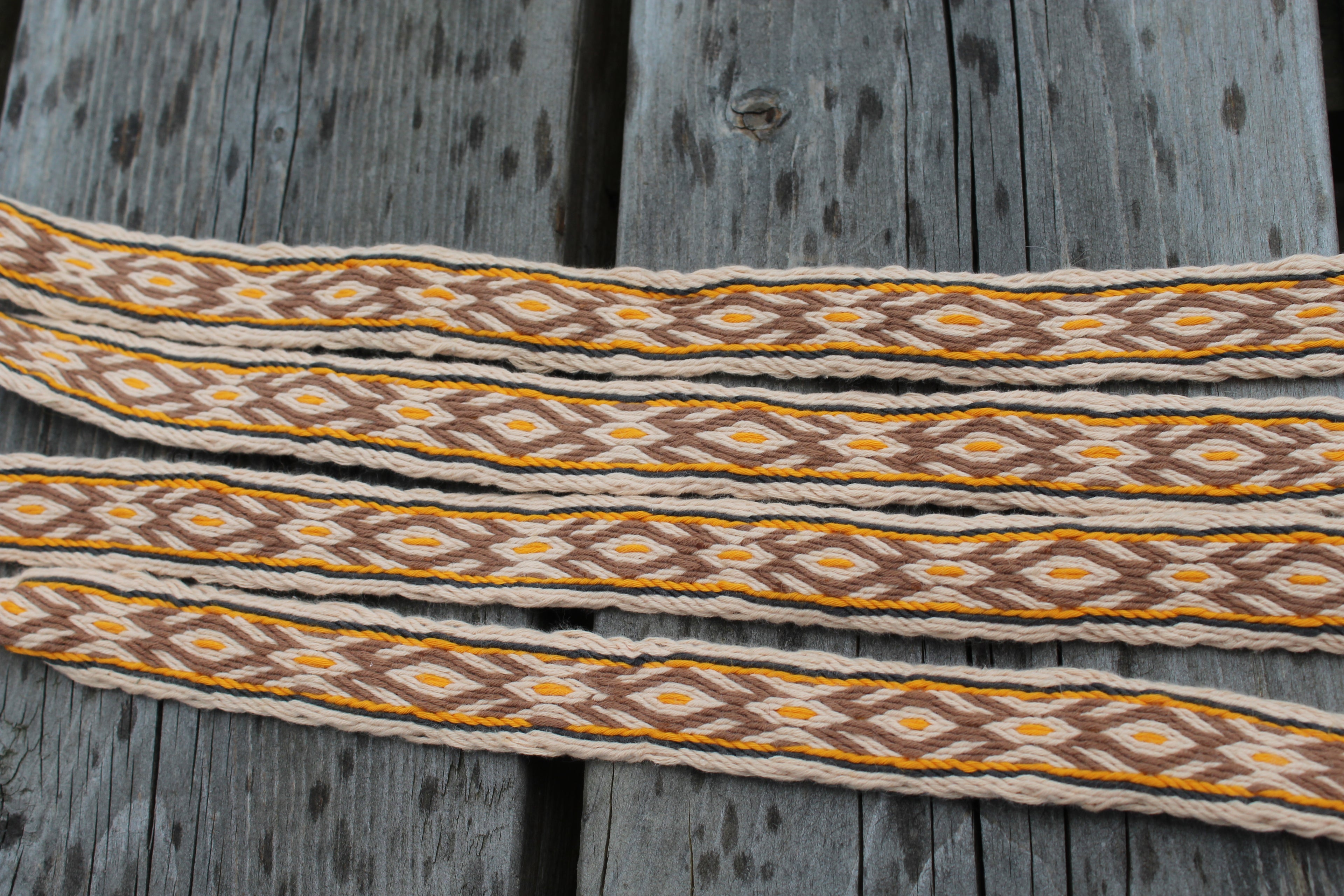 A close-up of a woven decorative strap with a geometric diamond pattern in brown, mustard yellow, and cream, laid flat on weathered gray wooden planks.
