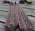 A woven shoulder strap or belt laid flat on weathered gray wooden planks, featuring a black, beige, and brown geometric pattern with a vibrant magenta-pink border.