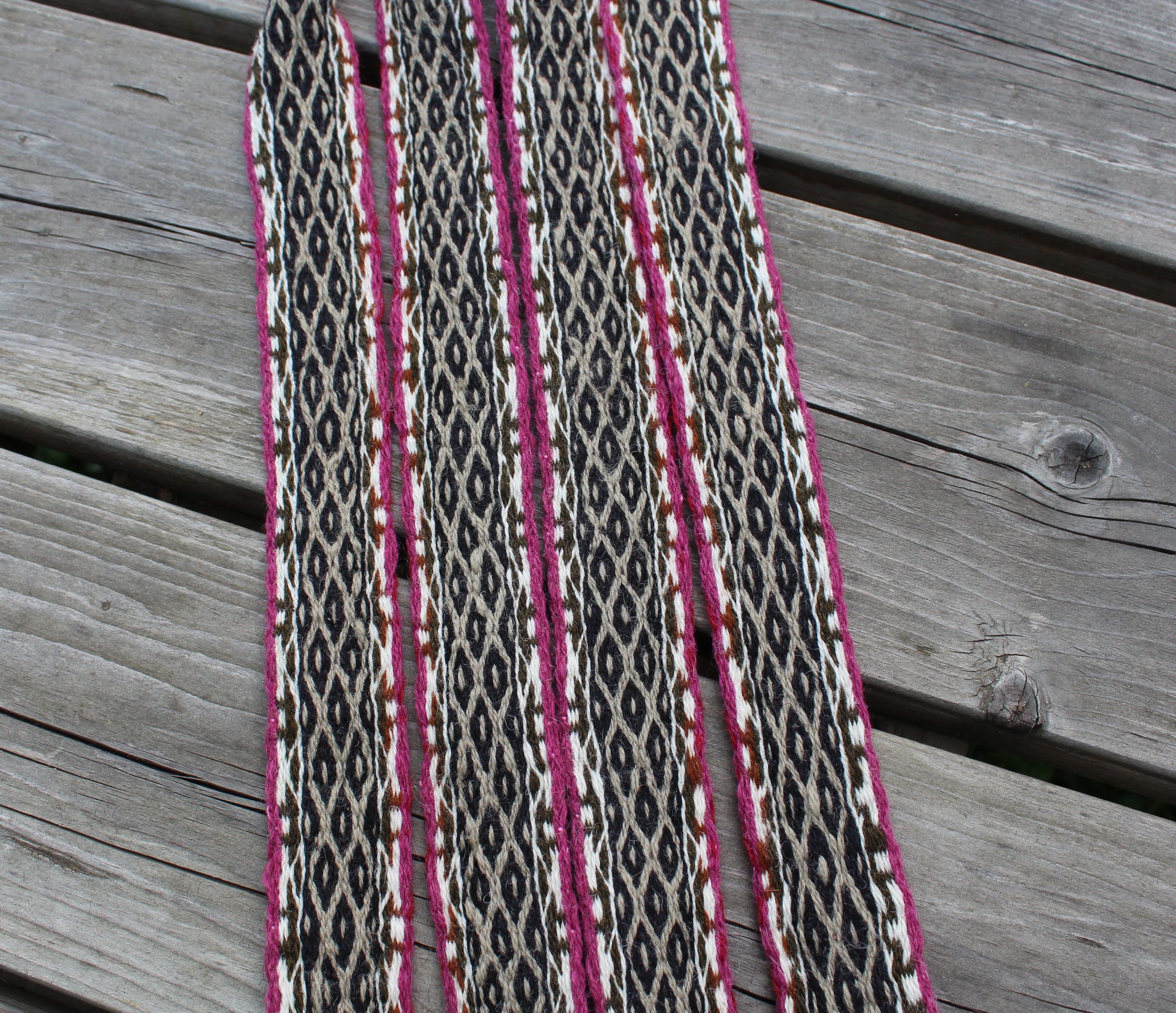 A long woven textile strip with an intricate black, white, and brown geometric pattern, accented by bright pink stripes, laid flat on weathered gray wooden planks.