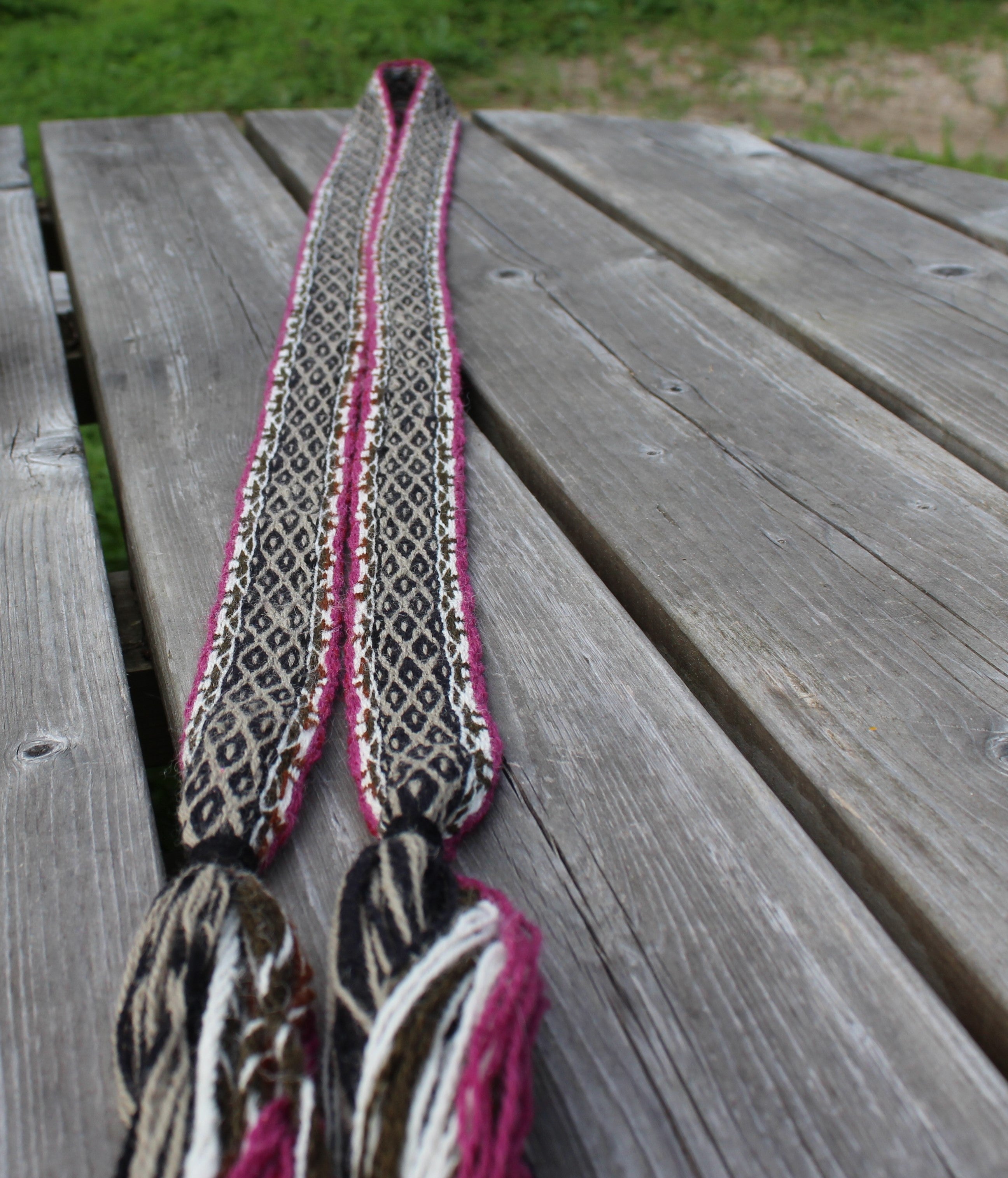 A long woven scarf with a black, white, and pink geometric pattern and tassel ends, laid out on a weathered gray wooden table.