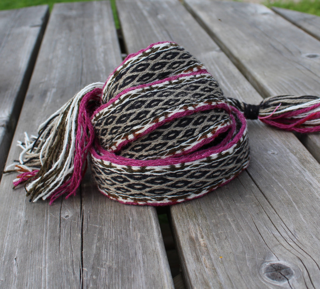 A woven scarf with a black, beige, and brown geometric pattern and magenta-pink tassels, resting on weathered gray wooden planks.