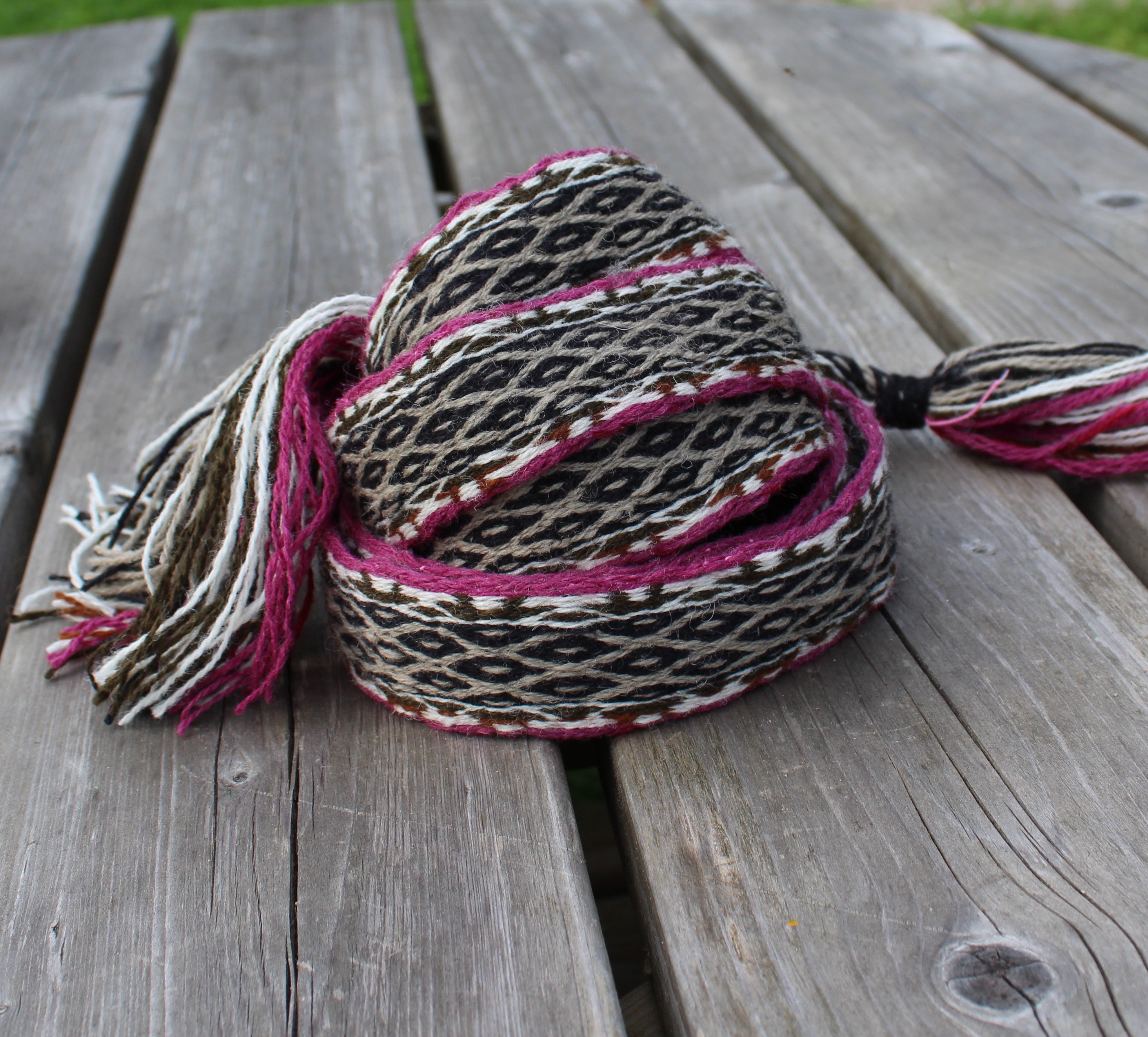 A woven scarf with a black, beige, and brown geometric pattern and magenta-pink tassels, resting on weathered gray wooden planks.