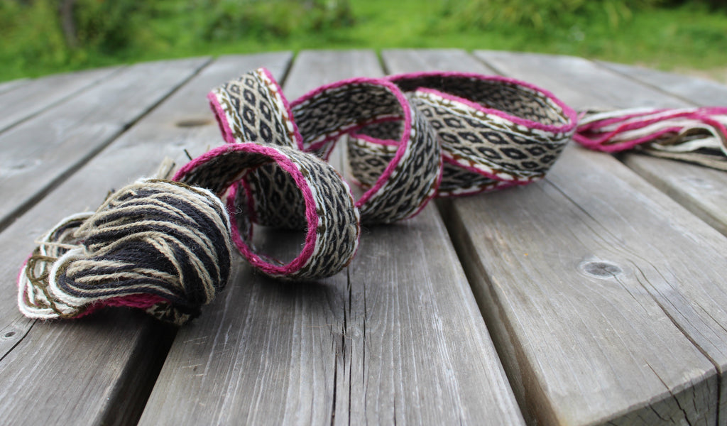 A handmade woven rope garland with black, beige, and pink patterns, displayed on a weathered wooden deck with a blurred green background.