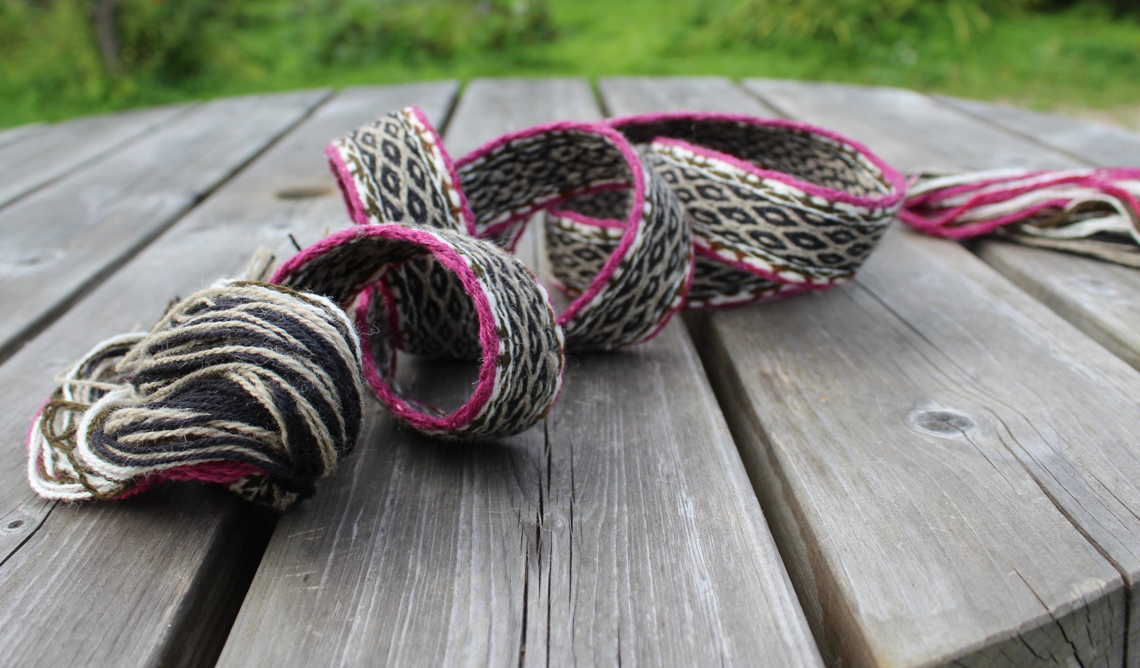 A handmade woven rope garland with black, beige, and pink patterns, displayed on a weathered wooden deck with a blurred green background.