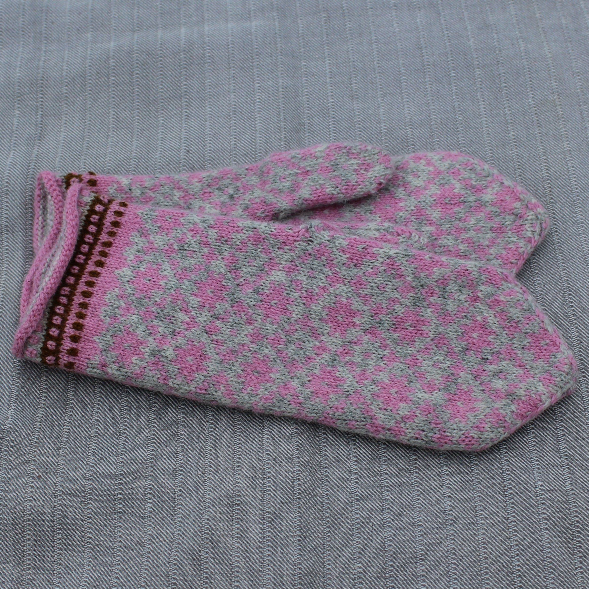 A single knit mitten glove with a pink and gray floral pattern and a brown decorative trim, laid flat on a gray textured fabric background.