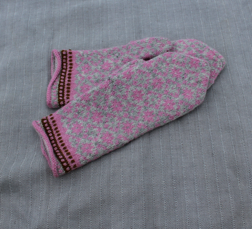 A pair of knit fingerless mittens in pink and gray with a floral pattern and brown trim, laid flat on a gray textured fabric background.