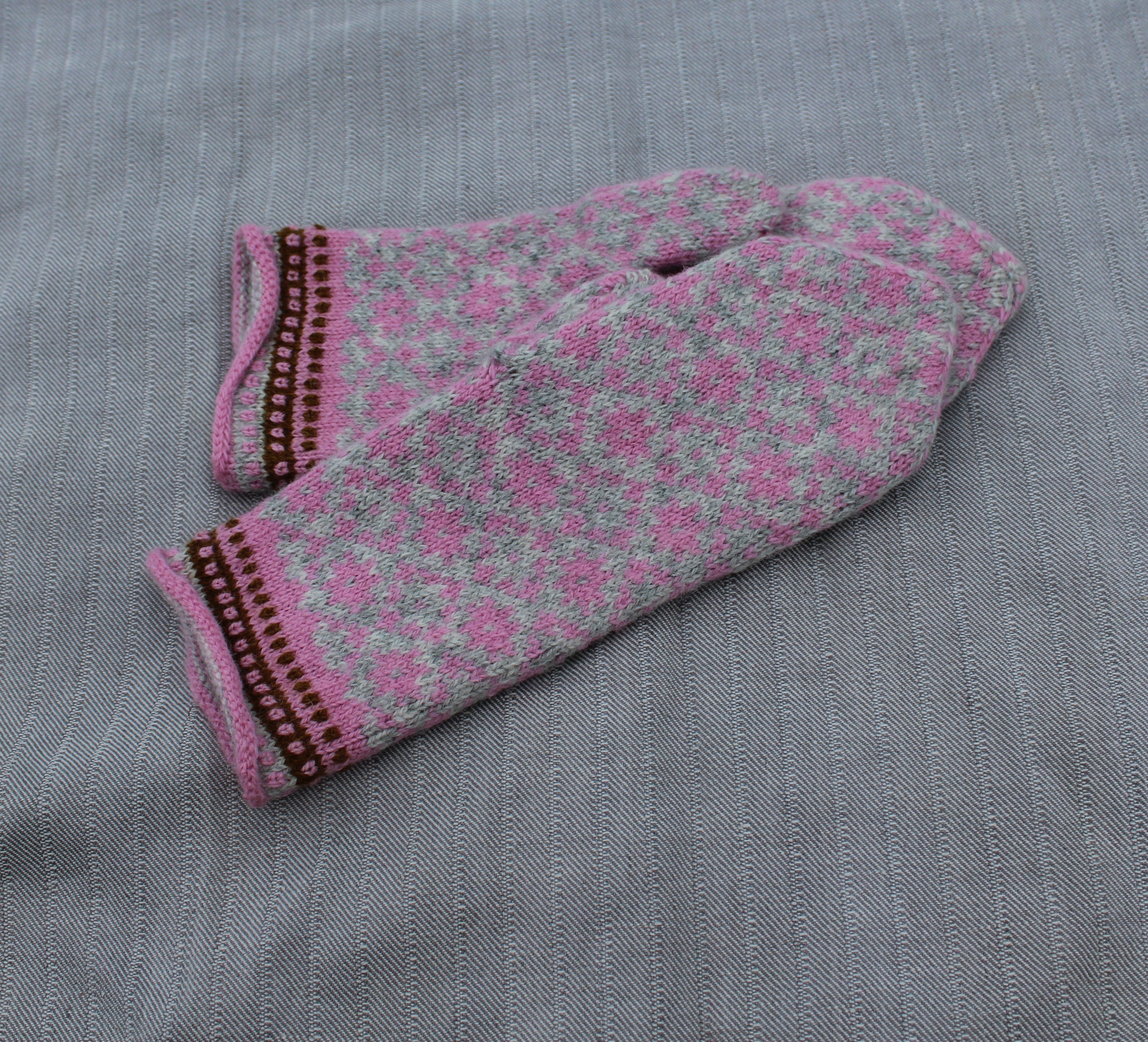 A pair of knit fingerless mittens in pink and gray with a floral pattern and brown trim, laid flat on a gray textured fabric background.