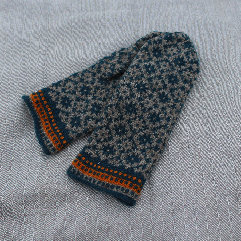 A pair of knit mittens with a dark teal and beige snowflake pattern and orange striped trim, laid flat on a light gray textured fabric background.