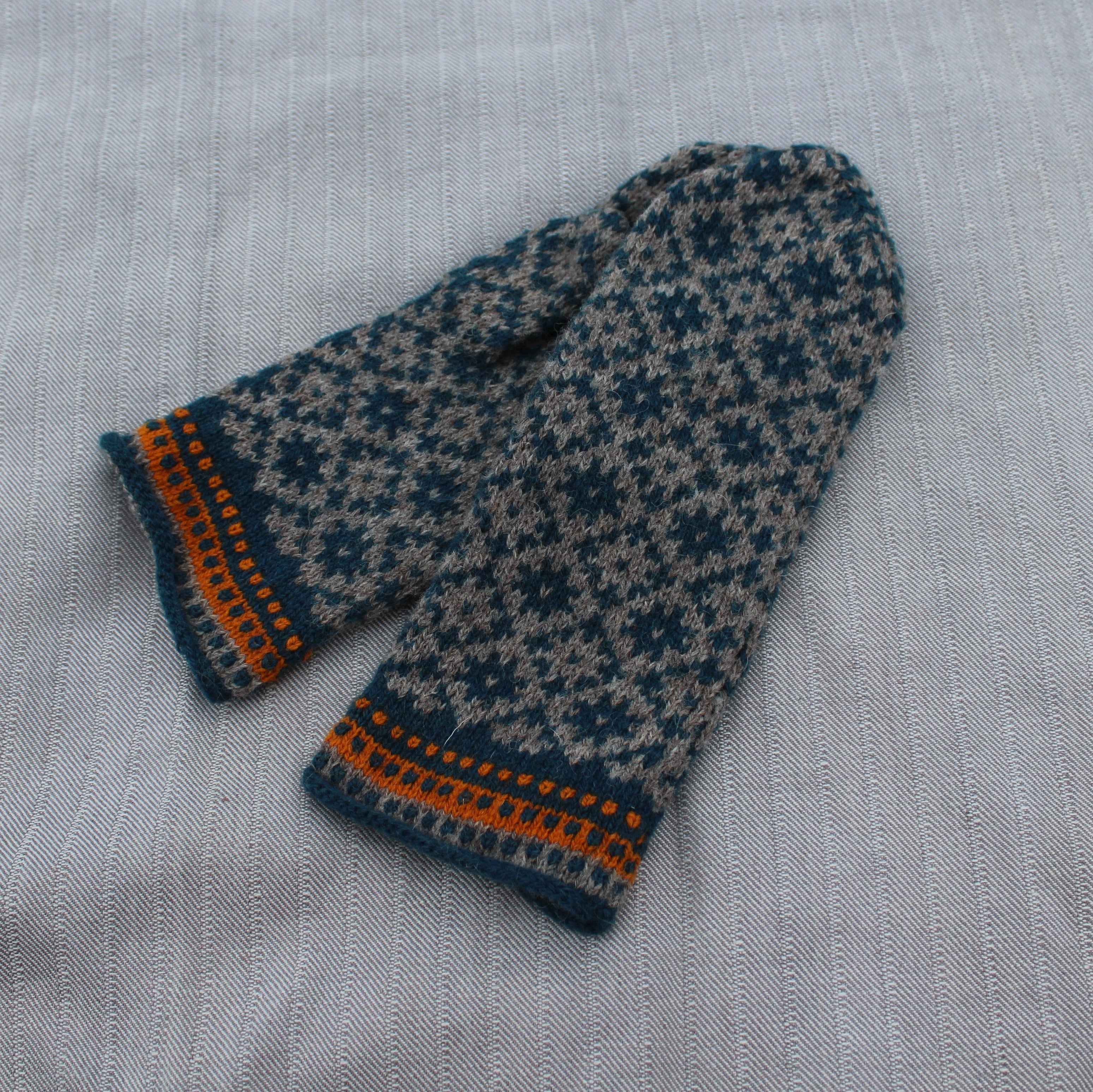 A pair of knit mittens with a dark teal and beige snowflake pattern and orange striped trim, laid flat on a light gray textured fabric background.
