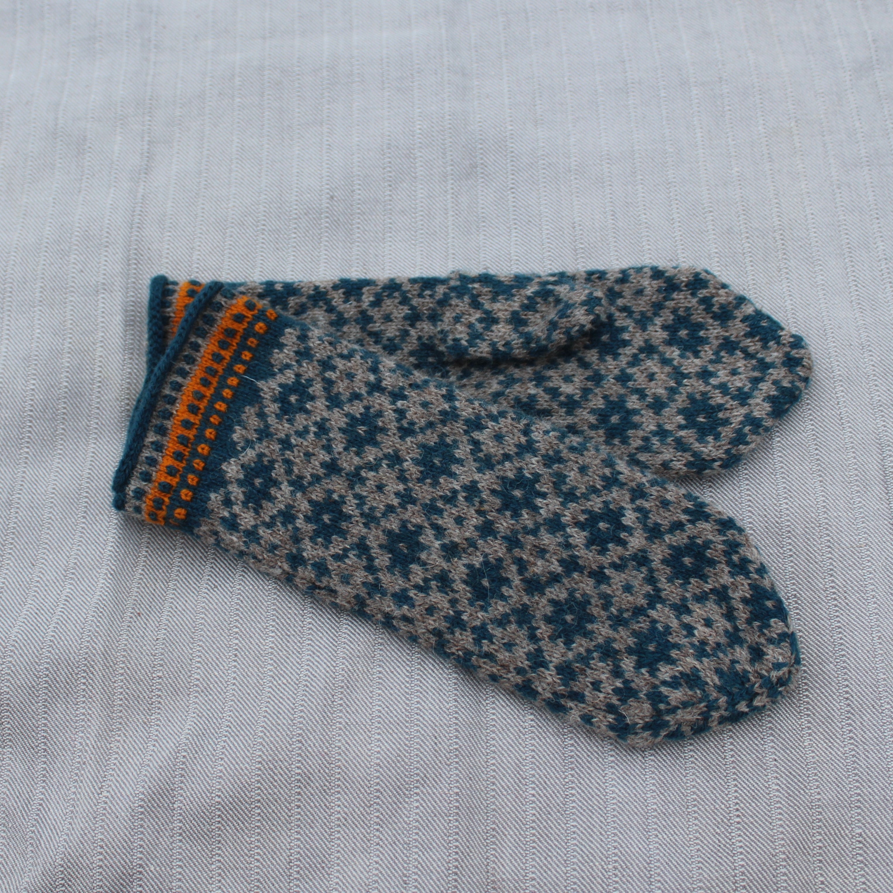 A single knit mitten with a dark teal and beige snowflake pattern, featuring a striped cuff with orange and dark teal stripes, laid flat on a light gray textured fabric background.