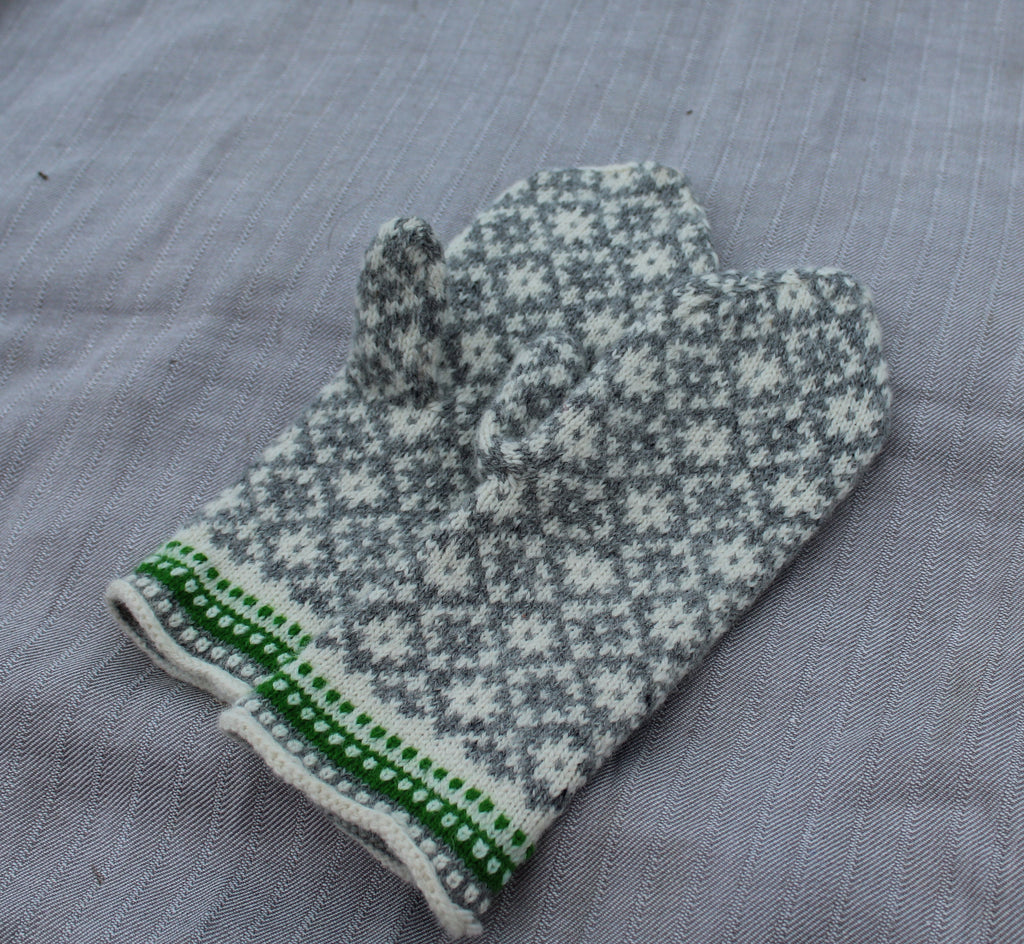 A single knit mitten in a gray and white Nordic snowflake pattern with a green and white striped cuff, laid flat on a gray textured fabric background.