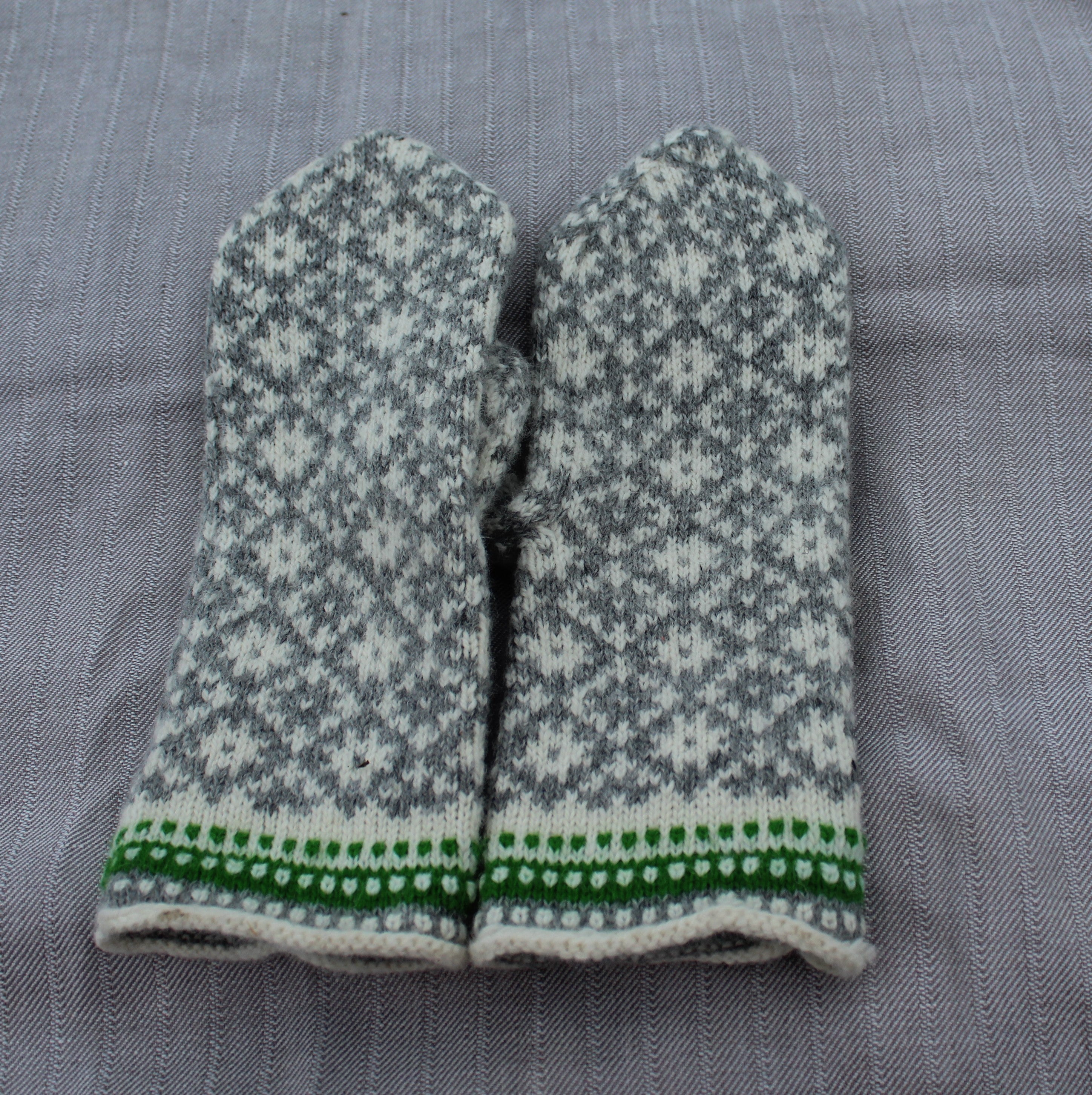 A pair of knit mittens with a gray and white Nordic snowflake pattern and a green and white patterned cuff, laid flat on a gray textured fabric background.