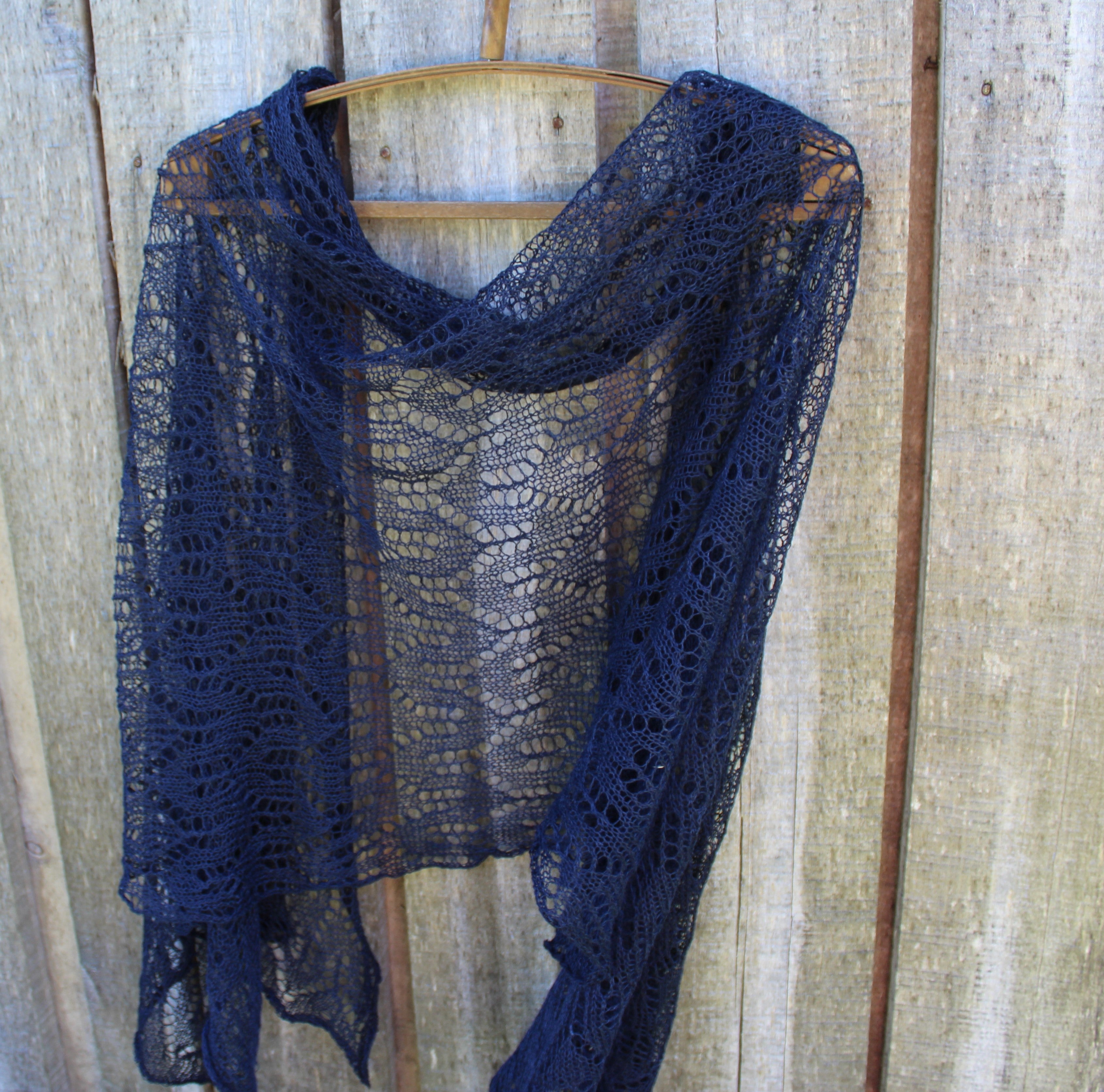 A navy blue open-knit lace shawl draped over a wooden hanger against a rustic light brown wooden plank wall.