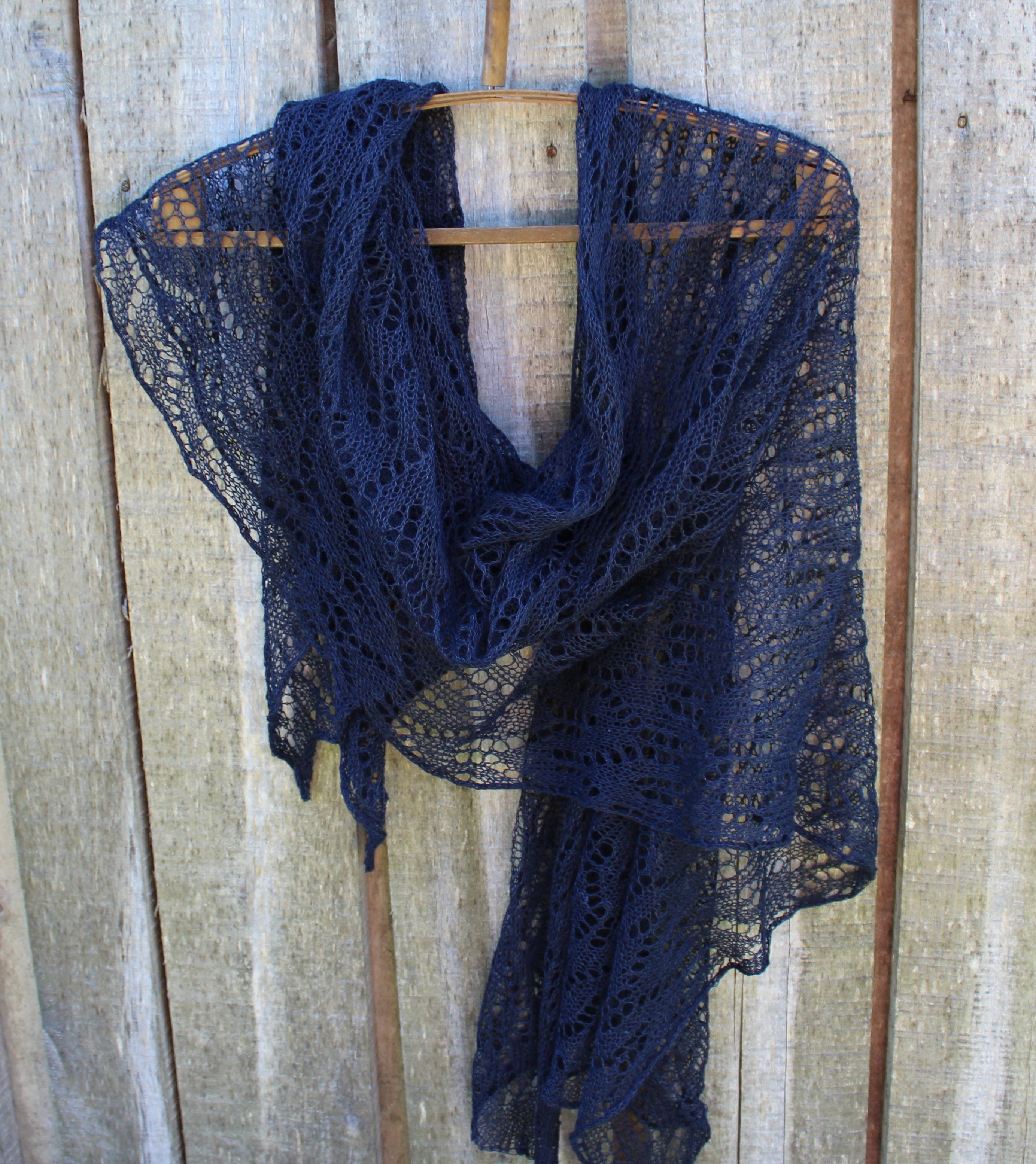 A navy blue open-knit lace shawl draped over a wooden hanger against a rustic light wood plank background.
