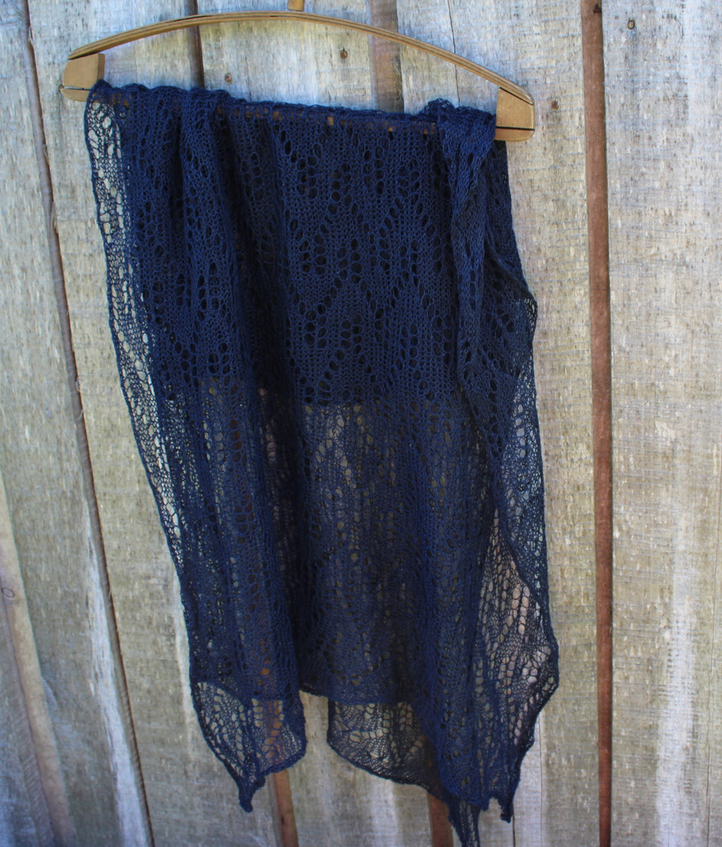 A dark navy blue open-knit lace shawl draped over a wooden hanger against a rustic wooden plank wall.
