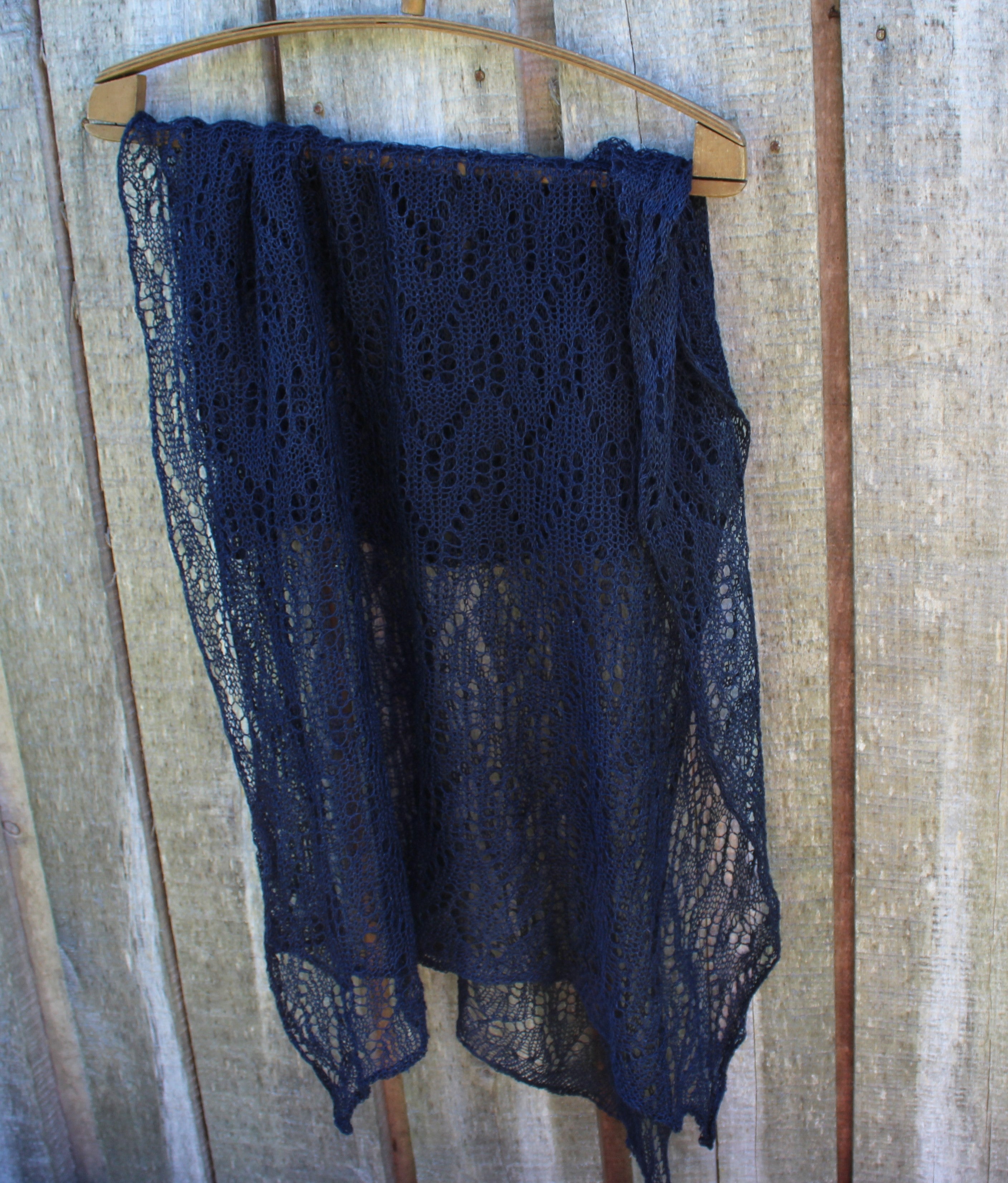A dark navy blue open-knit lace shawl draped over a wooden hanger against a rustic wooden plank wall.