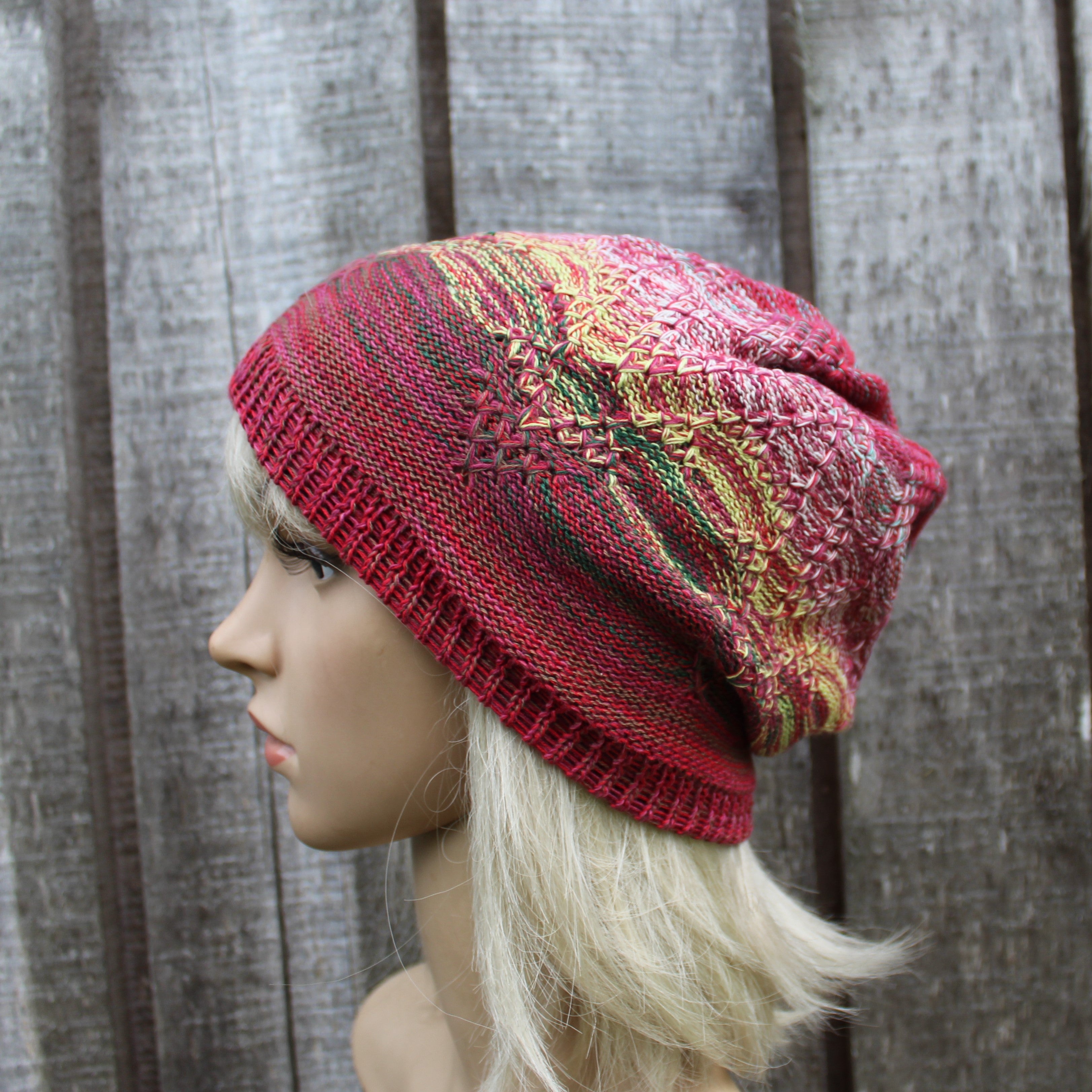 A multicolor knit slouchy hat with a ribbed brim, shown on a blonde mannequin head against a gray wooden plank background.