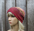 A close-up side view of a mannequin head wearing a knitted slouchy beanie hat. The hat is primarily deep red with a multicolor variegated pattern on the side, set against a rustic gray wooden fence background.