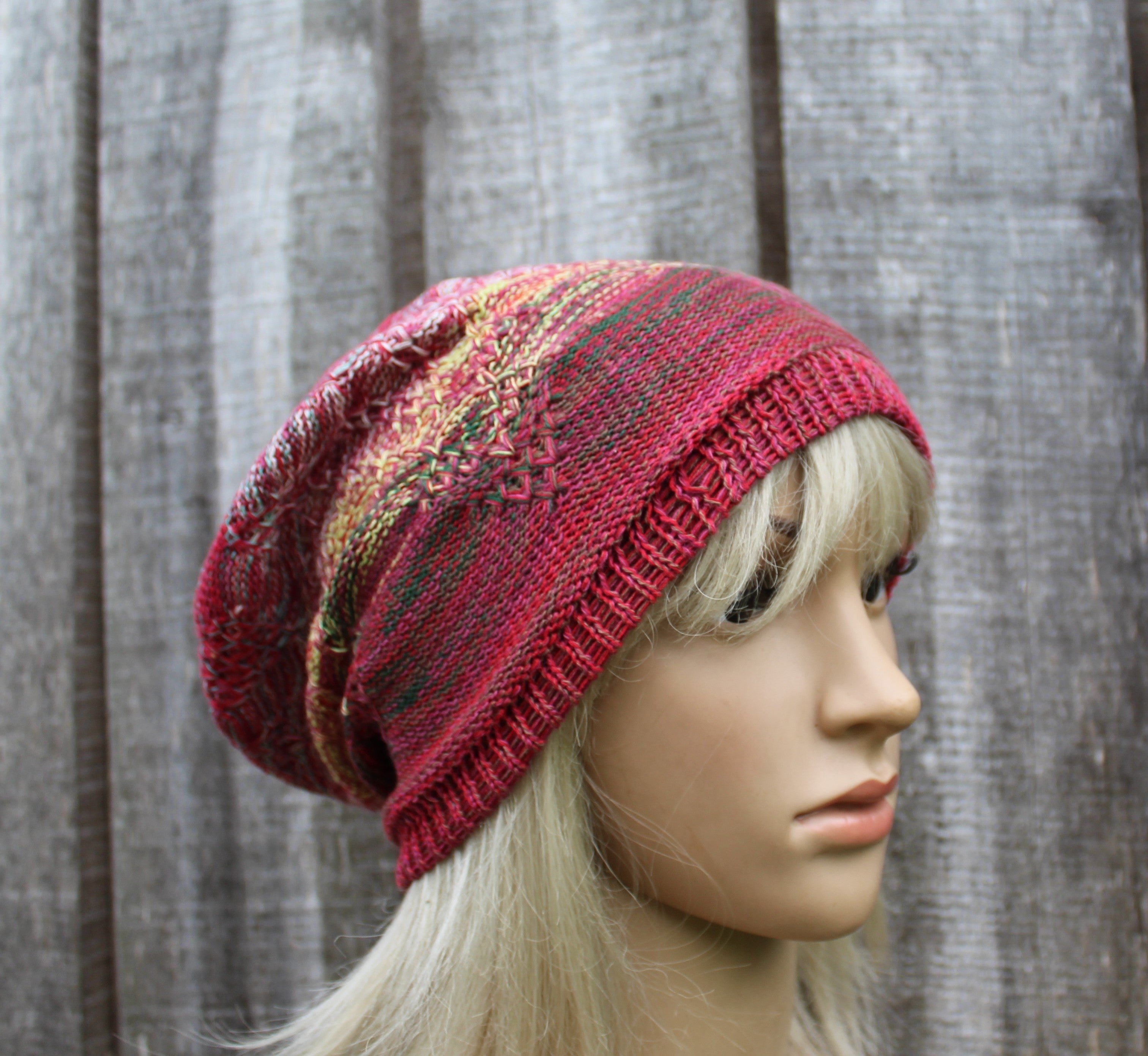 A knitted slouchy hat in a heathered red, pink, and green pattern, shown on a blonde mannequin head against a gray wooden plank background.