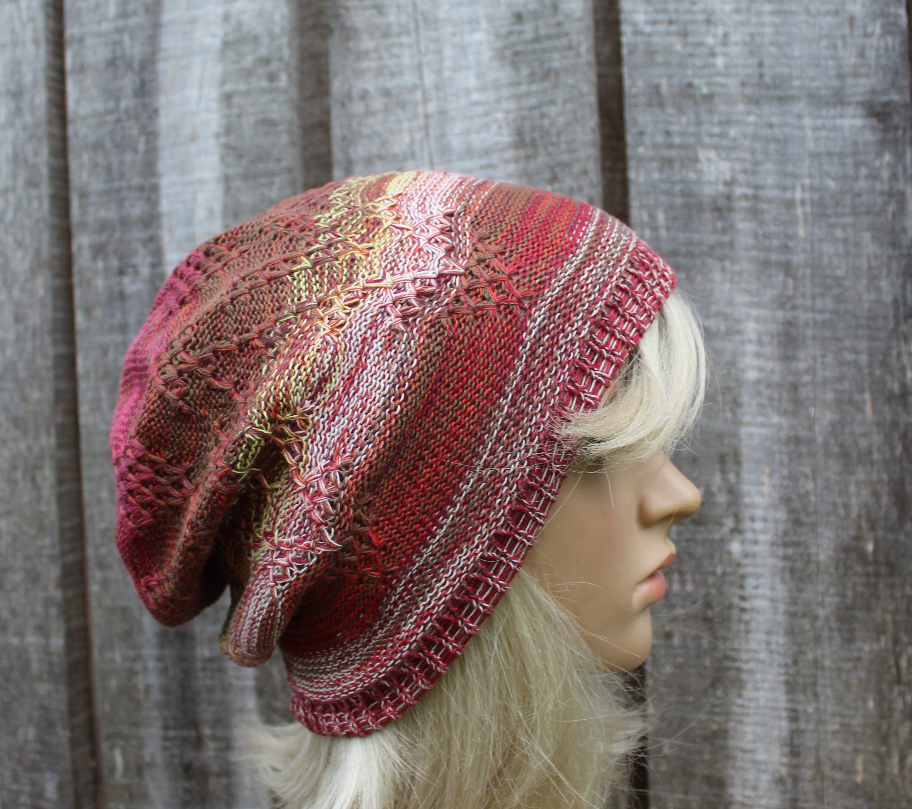 A knitted slouchy beanie hat in shades of pink, red, and beige with a textured pattern, displayed on a blonde mannequin head against a gray wooden plank background.