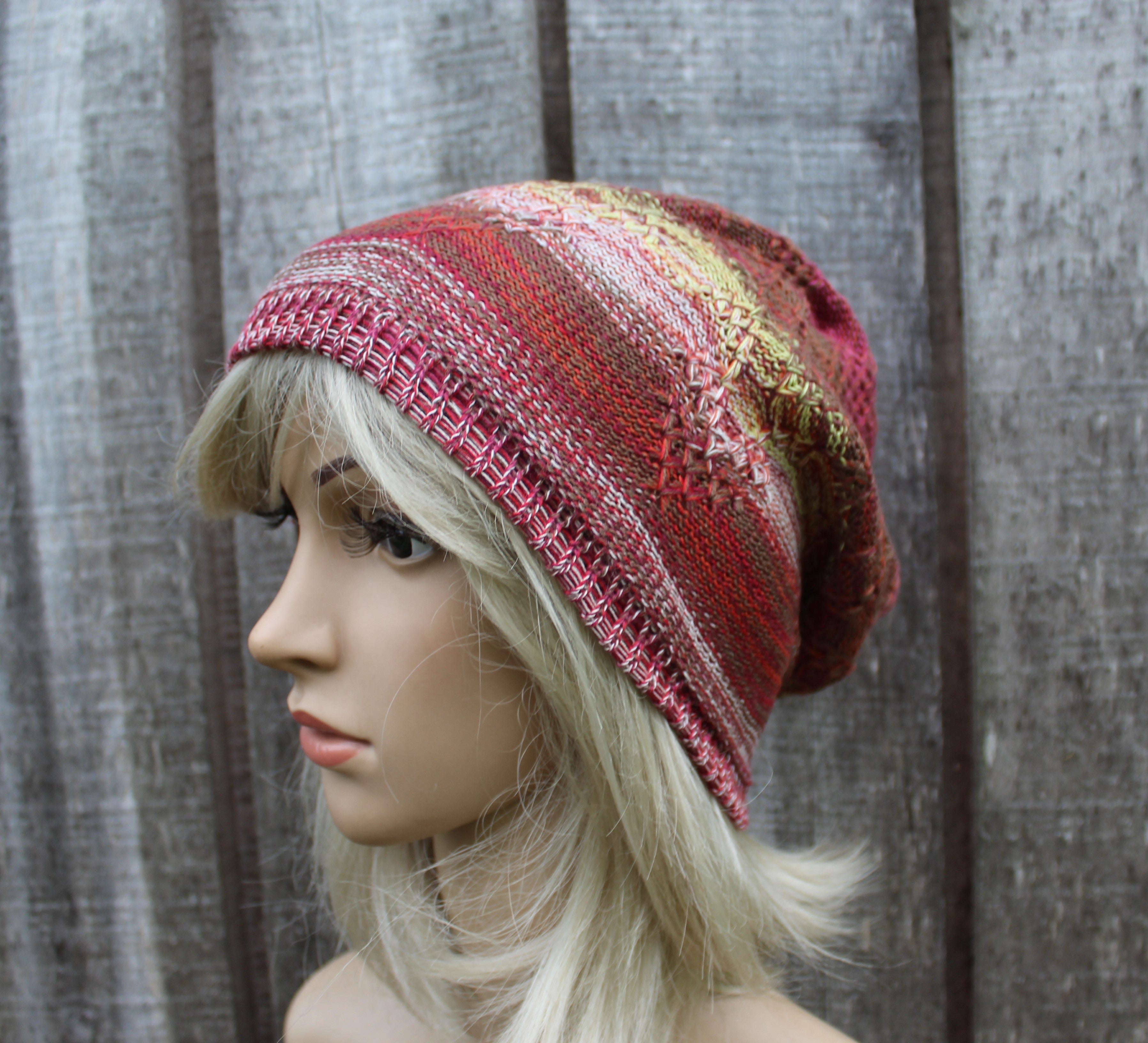 A multicolor striped knit slouchy beanie hat in shades of red, pink, orange, and gold, displayed on a blonde mannequin head against a gray wooden plank background.