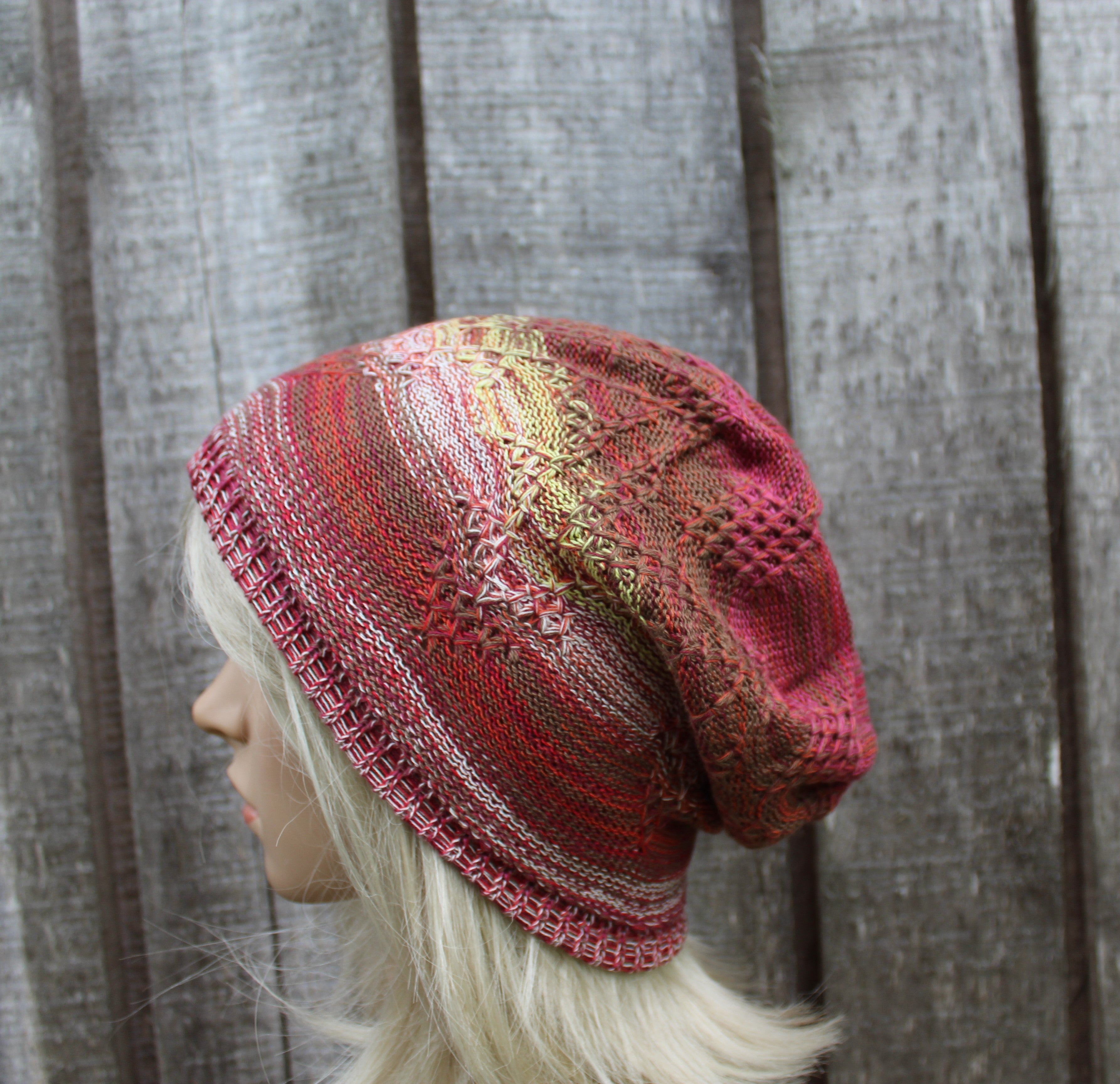 A knitted slouchy hat in shades of red, pink, and beige with a textured pattern, displayed on a blonde mannequin head against a gray wooden plank background.