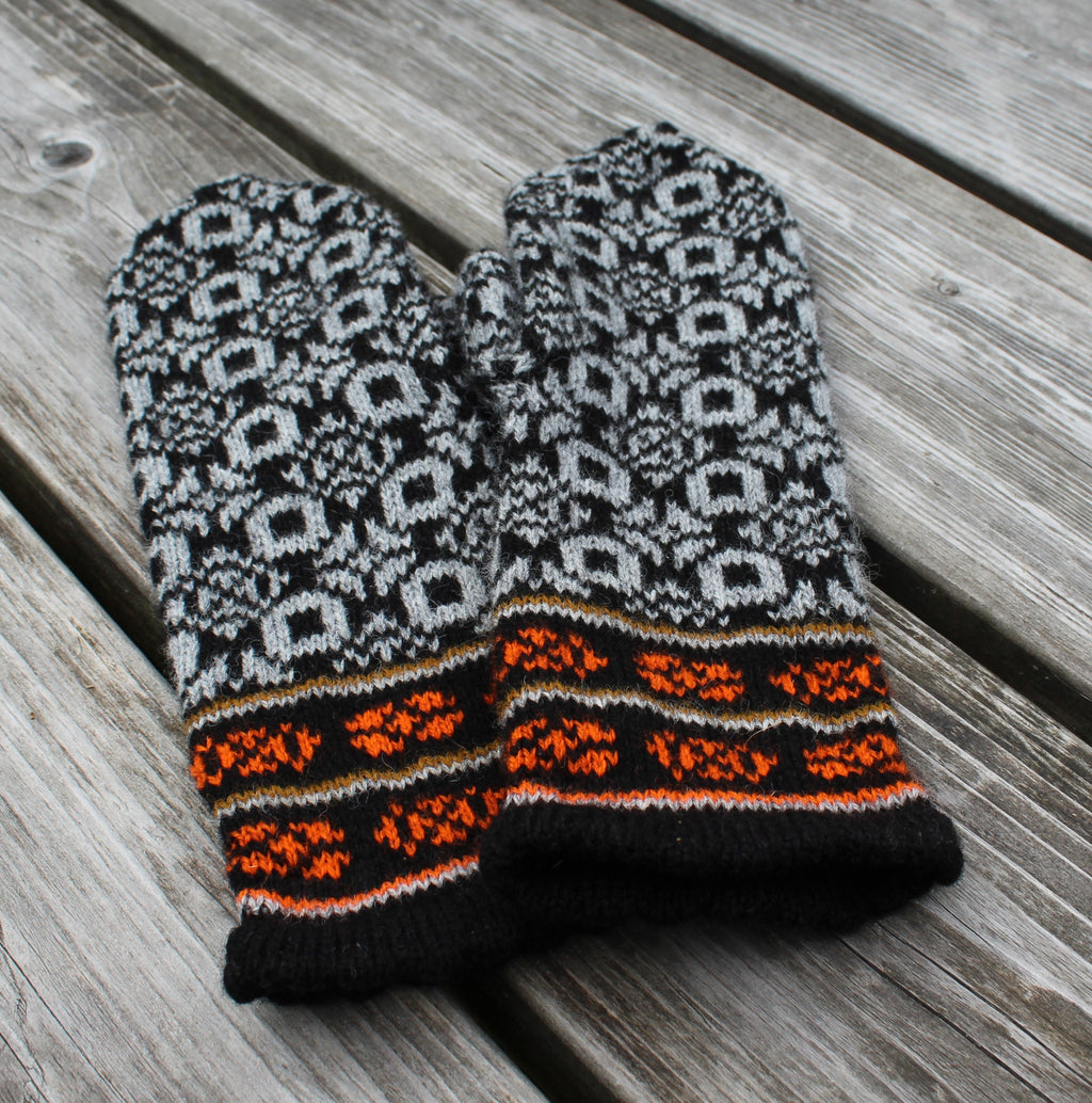 A pair of knit winter mittens with a black and gray Nordic snowflake pattern and bright orange accents, laid flat on weathered gray wooden planks.