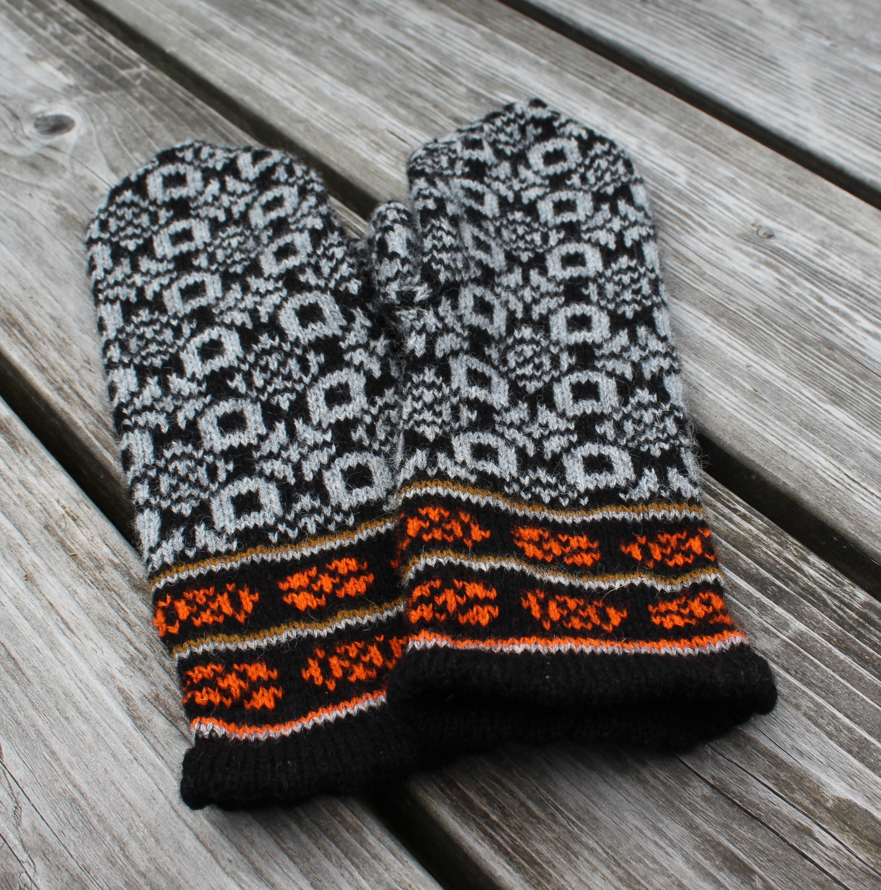 A pair of knit winter mittens with a black and gray Nordic snowflake pattern and bright orange accents, laid flat on weathered gray wooden planks.