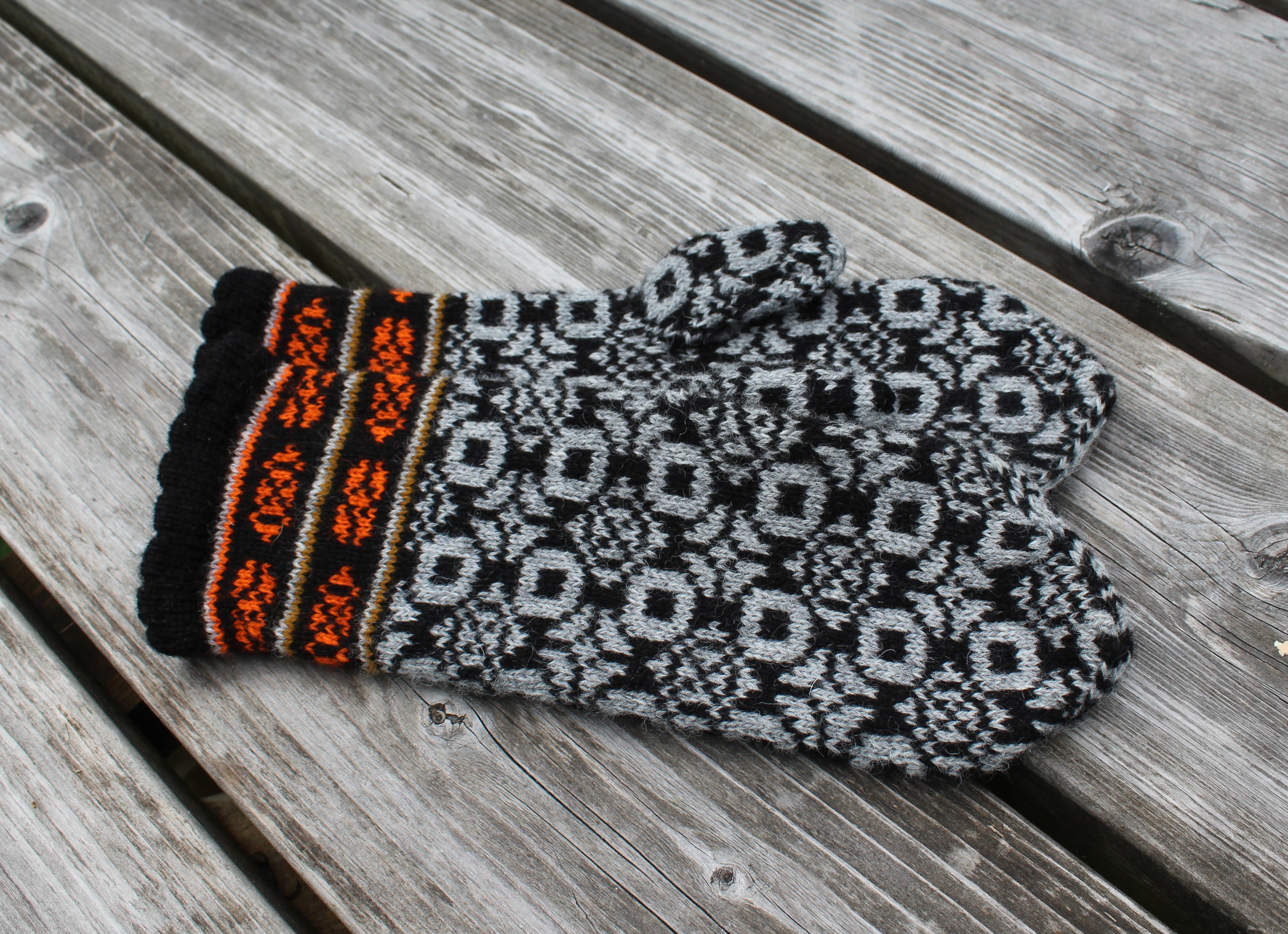 A single knit mitten-style glove with a black and gray geometric pattern and a striped cuff in black, orange, beige, and gray, placed on weathered wooden planks.