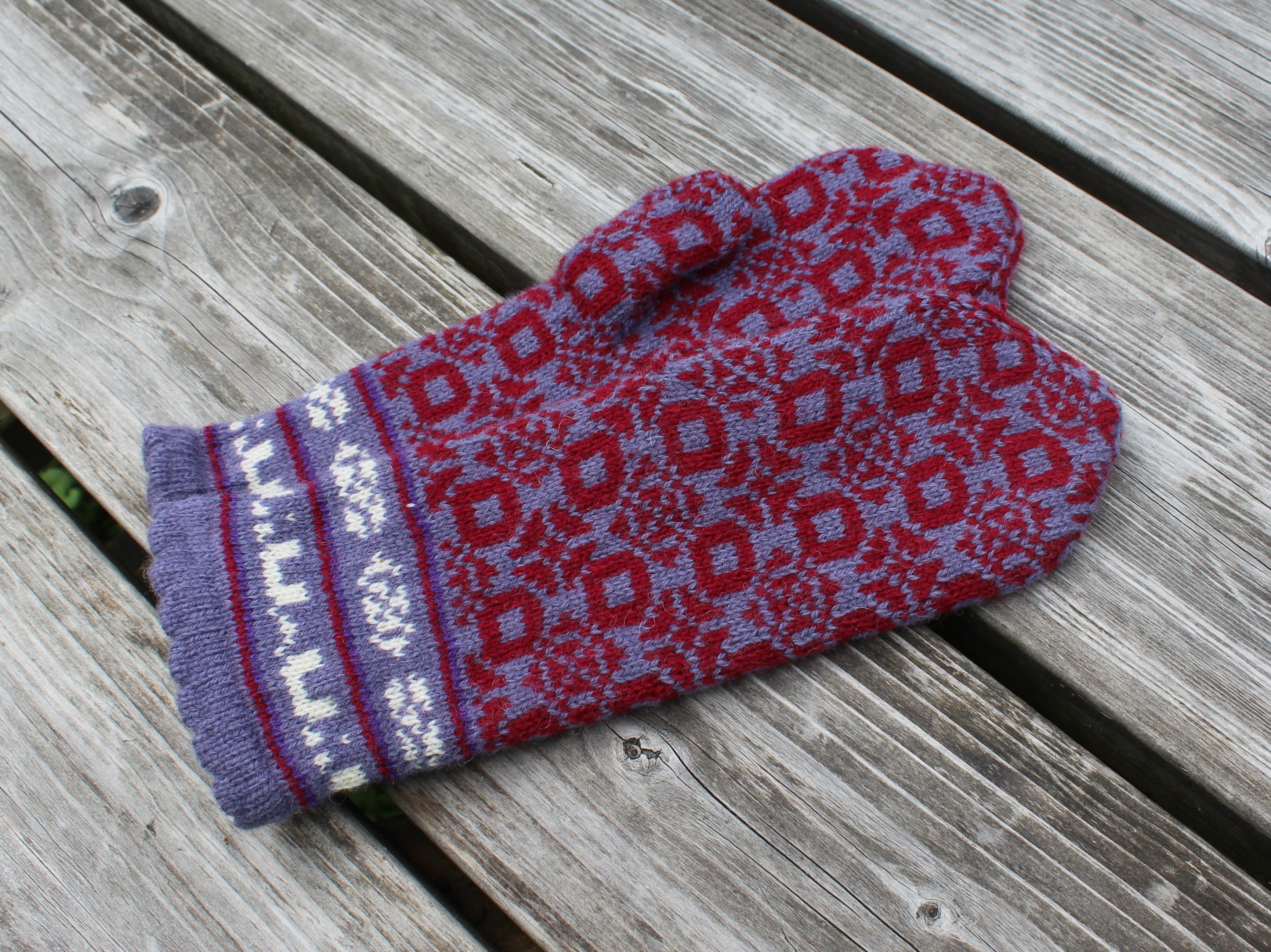 A single knit mitten with a red and purple geometric pattern and a purple cuff with white snowflake details, resting on weathered gray wooden planks.