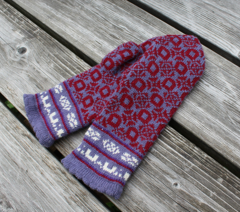 A pair of knit winter gloves with a red and purple geometric pattern and white patterned cuffs, displayed on a weathered gray wooden plank surface.
