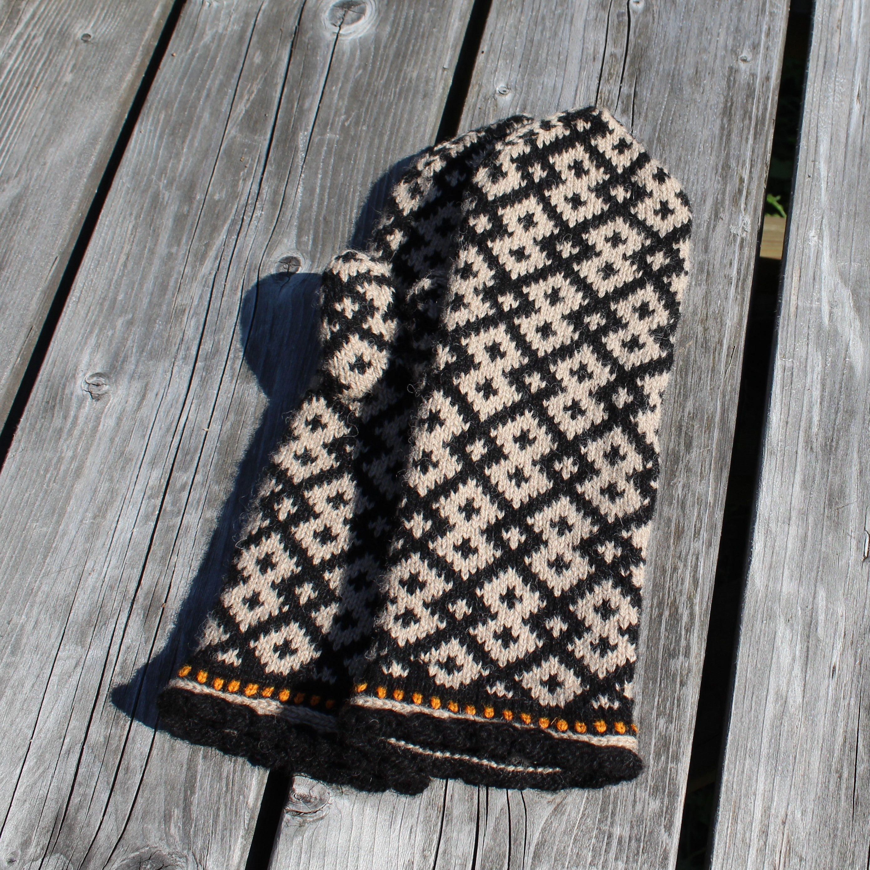 A hand-knit pet sweater or hat with a black and cream geometric diamond pattern, featuring a decorative orange and black striped trim, displayed on weathered gray wooden planks.