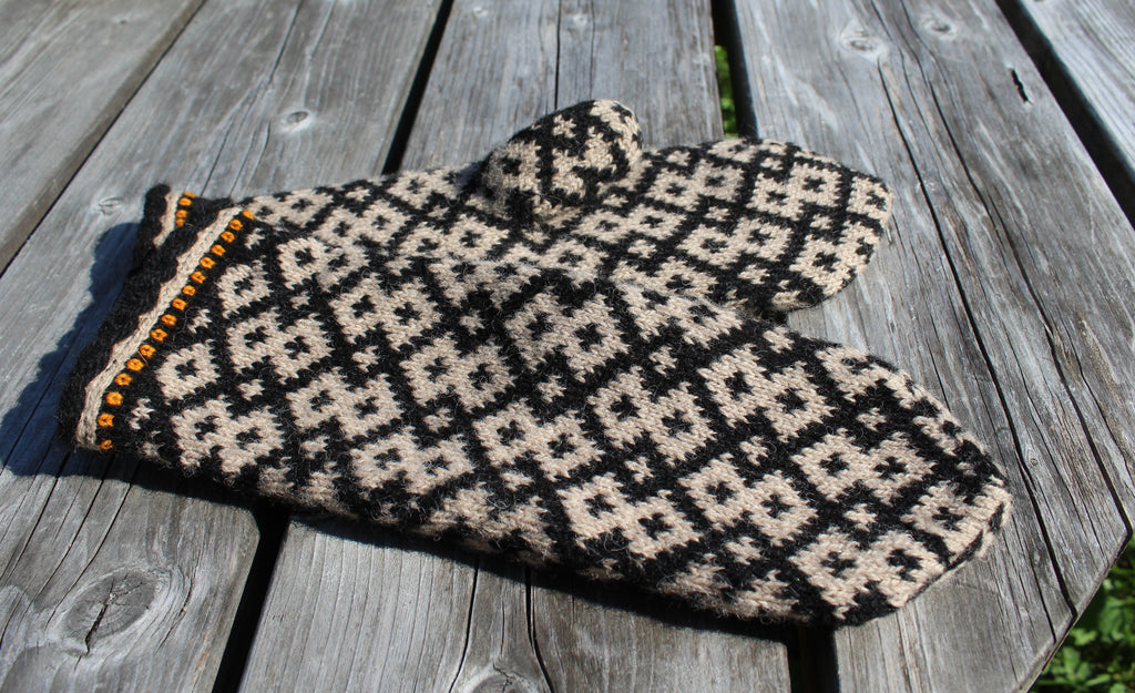 A pair of knit mittens with a black and beige geometric pattern, featuring a decorative orange and black trim on one edge, displayed on a weathered gray wooden surface.