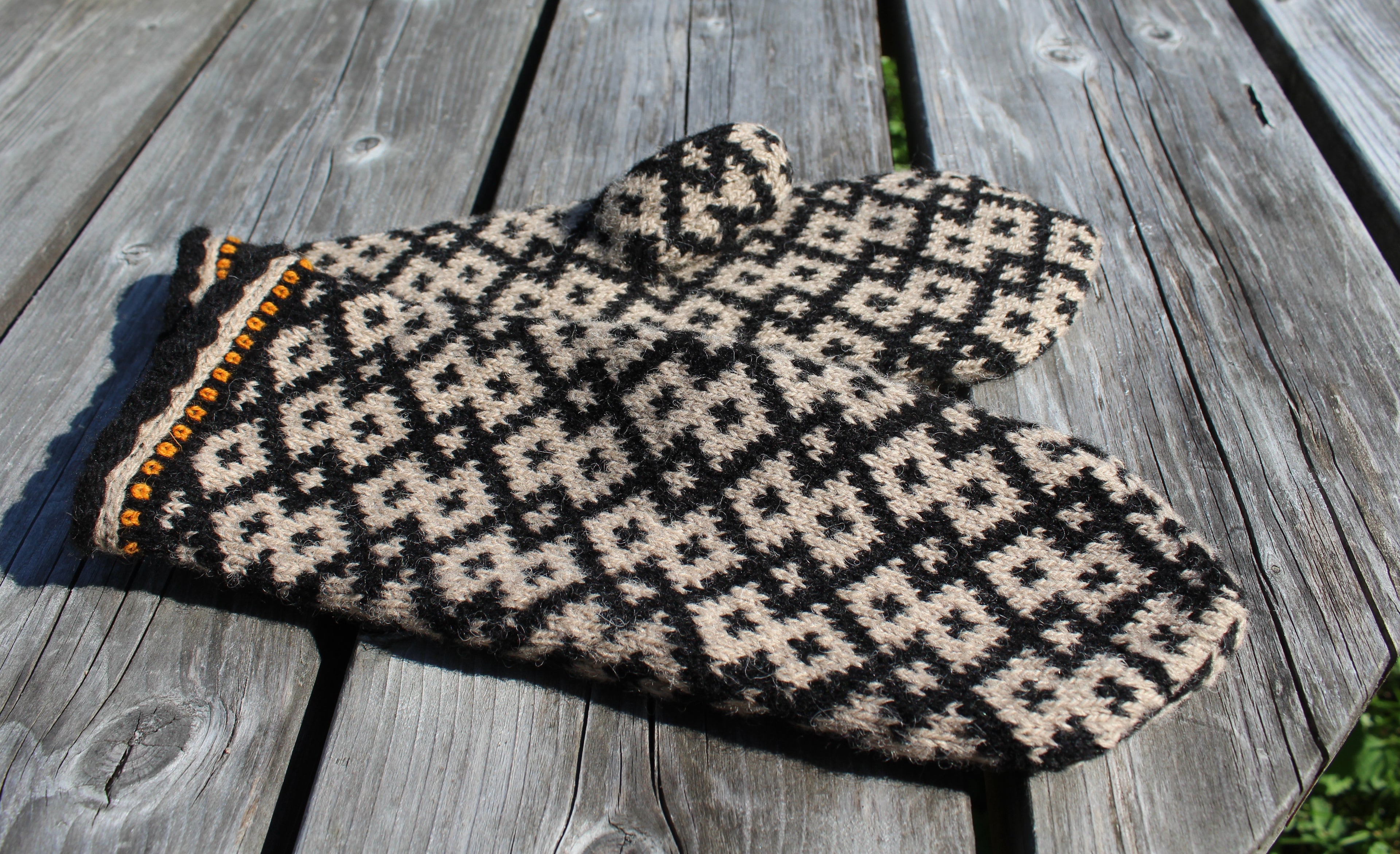 A pair of knit mittens with a black and beige geometric pattern, featuring a decorative orange and black trim on one edge, displayed on a weathered gray wooden surface.