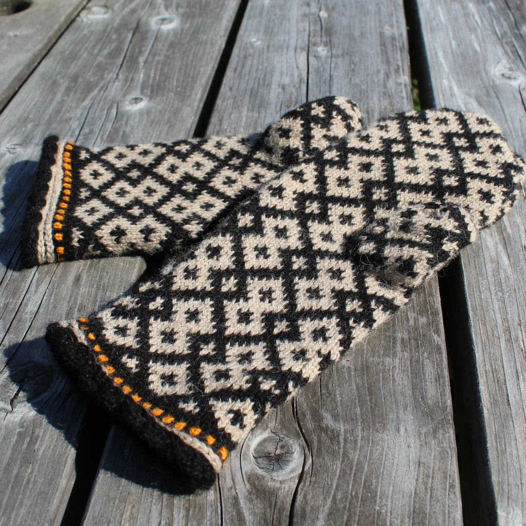 A pair of knit mittens with a black and beige geometric diamond pattern and orange trim, displayed on a weathered gray wooden deck.