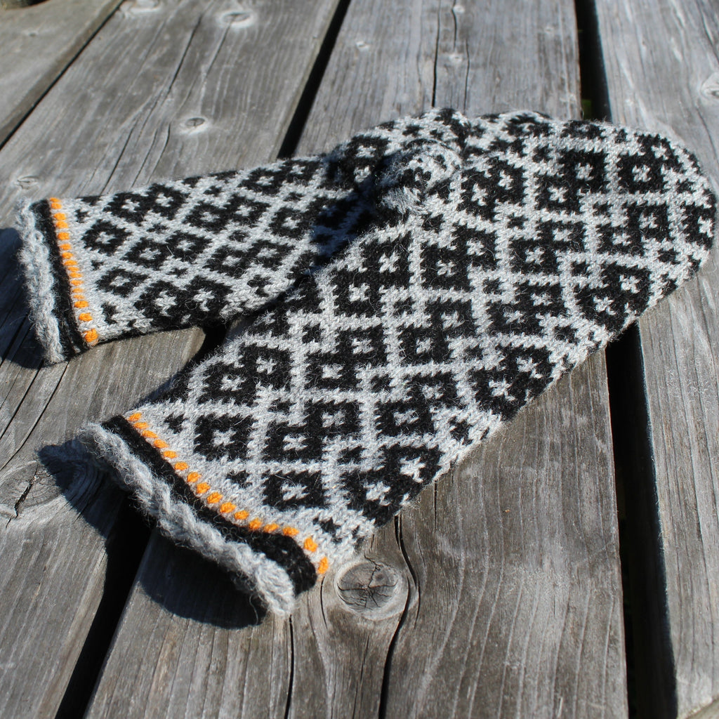 A pair of hand-knit mittens with a black and white geometric diamond pattern and orange stripe accents, displayed on weathered gray wooden planks.