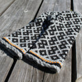 A knit winter hat with a black and white geometric pattern and a gray braided cuff, featuring a thin orange stripe detail, displayed on weathered wooden planks.