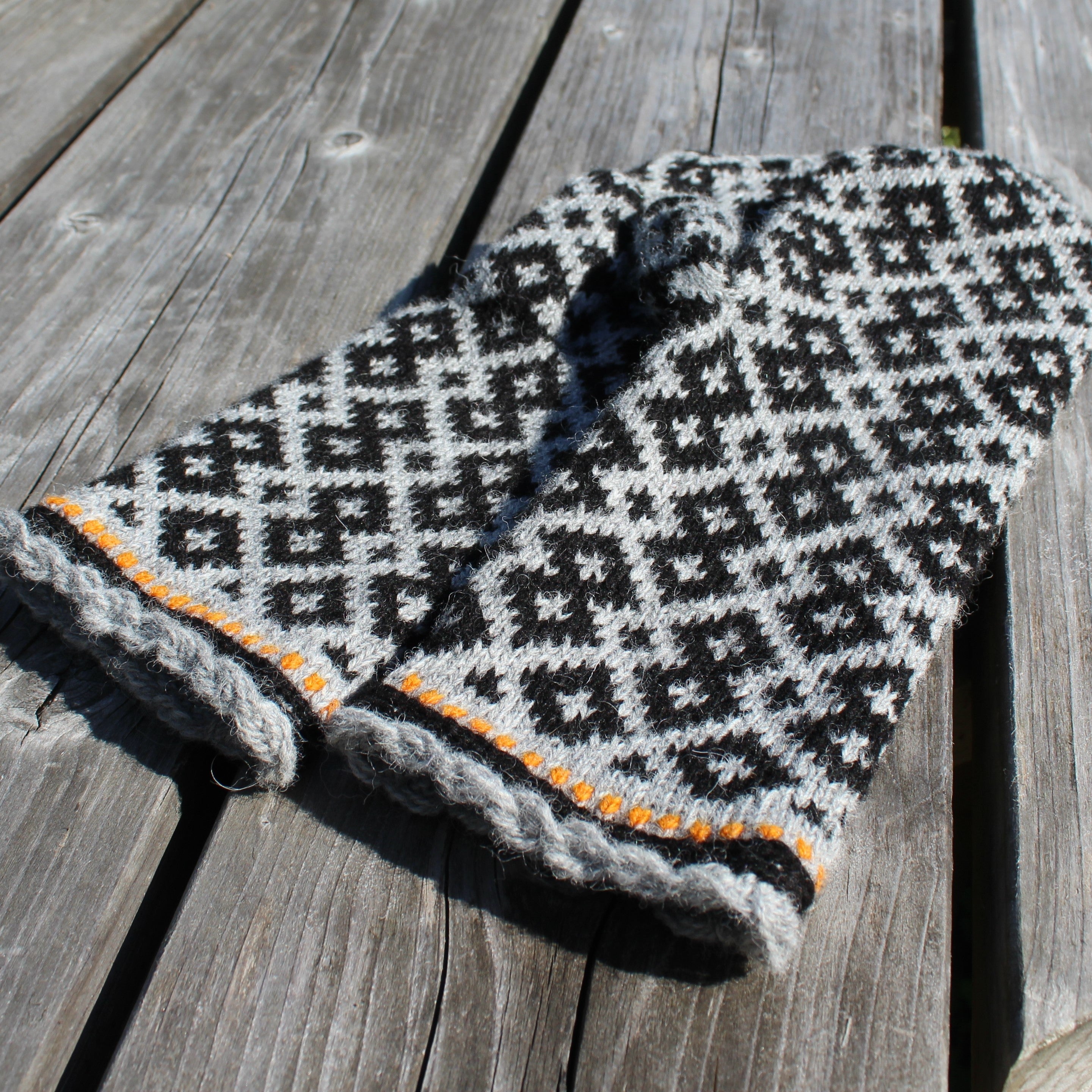 A knit winter hat with a black and white geometric pattern and a gray braided cuff, featuring a thin orange stripe detail, displayed on weathered wooden planks.