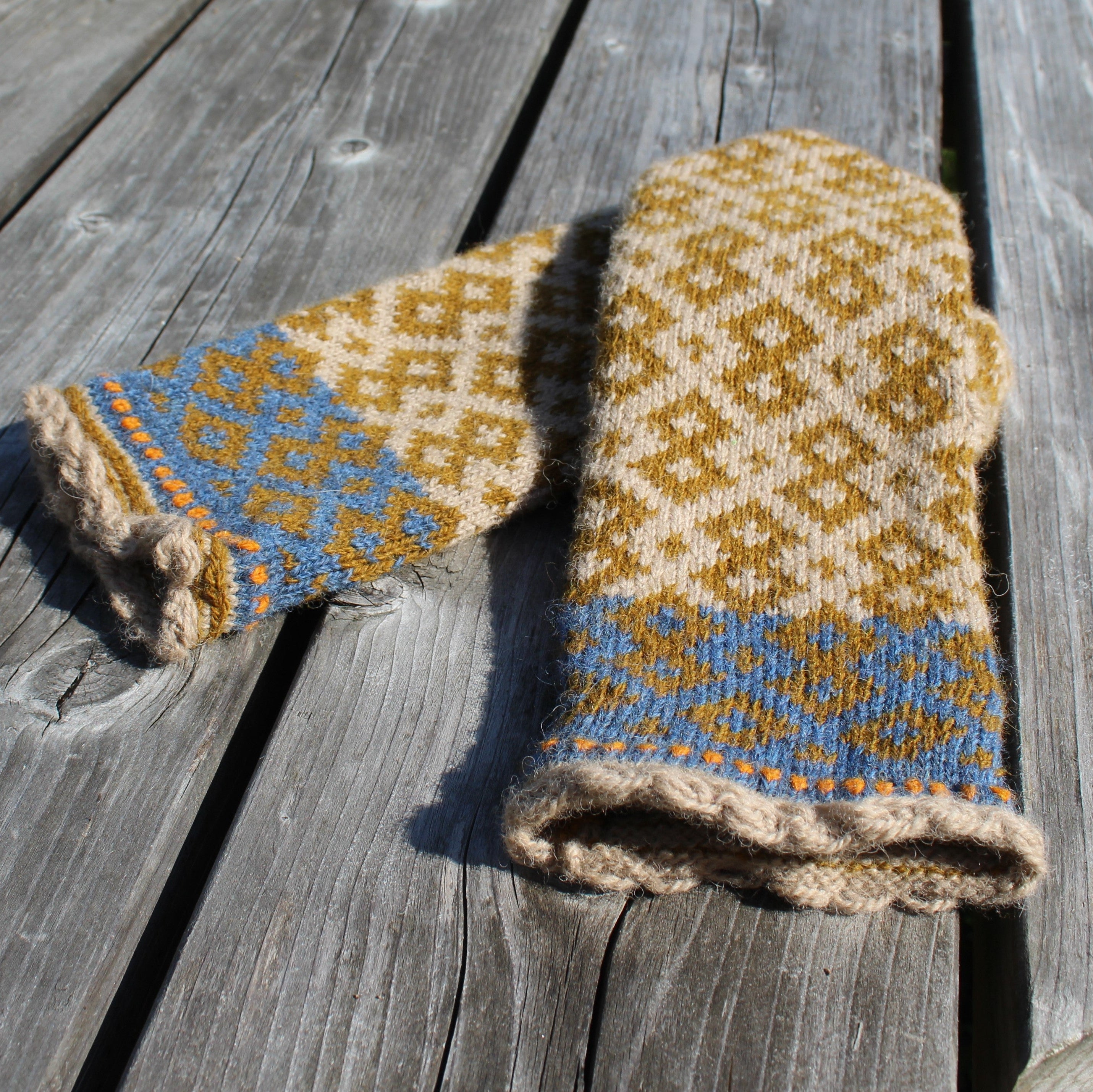 A pair of hand-knit wool mittens with a geometric pattern in mustard yellow, cream, and light blue, laid out on weathered gray wooden planks.