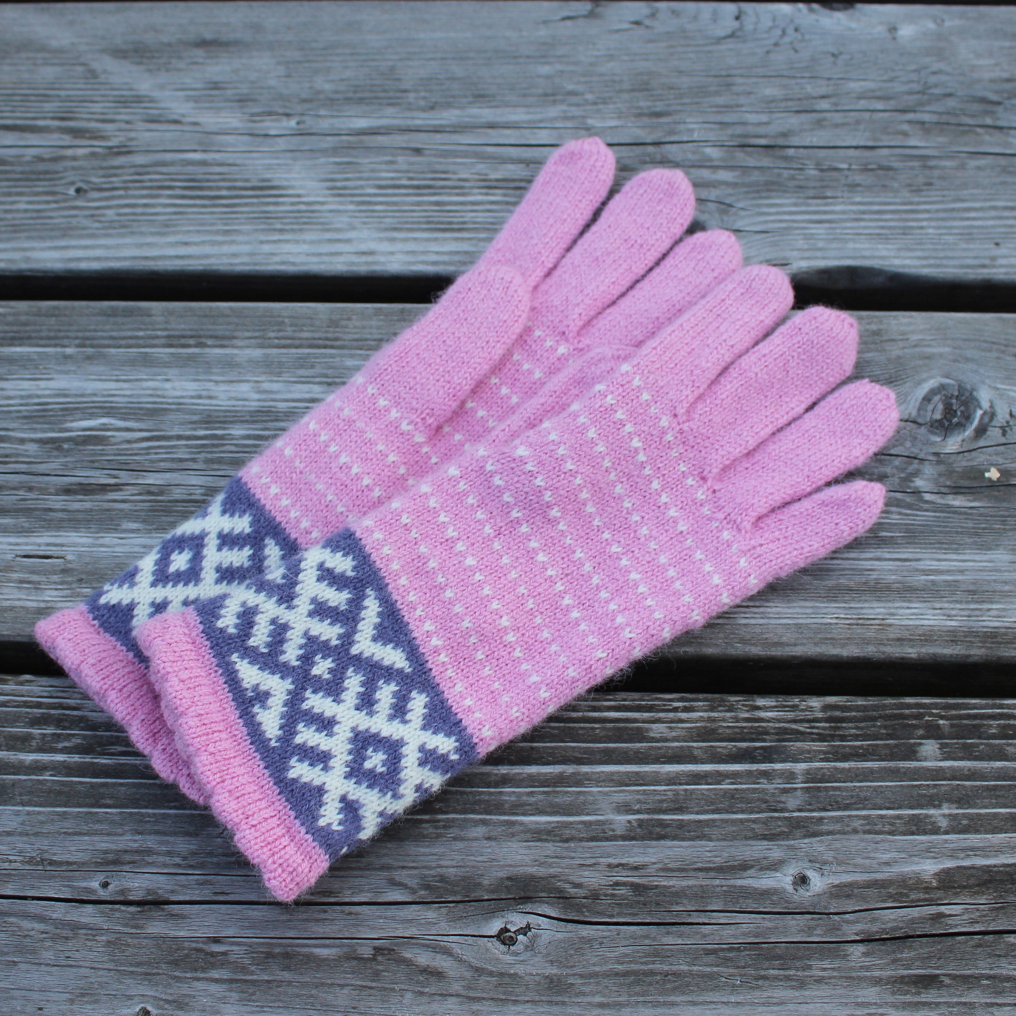 A pair of pink knit gloves with a white snowflake pattern on the cuff, displayed on a weathered gray wooden plank surface.